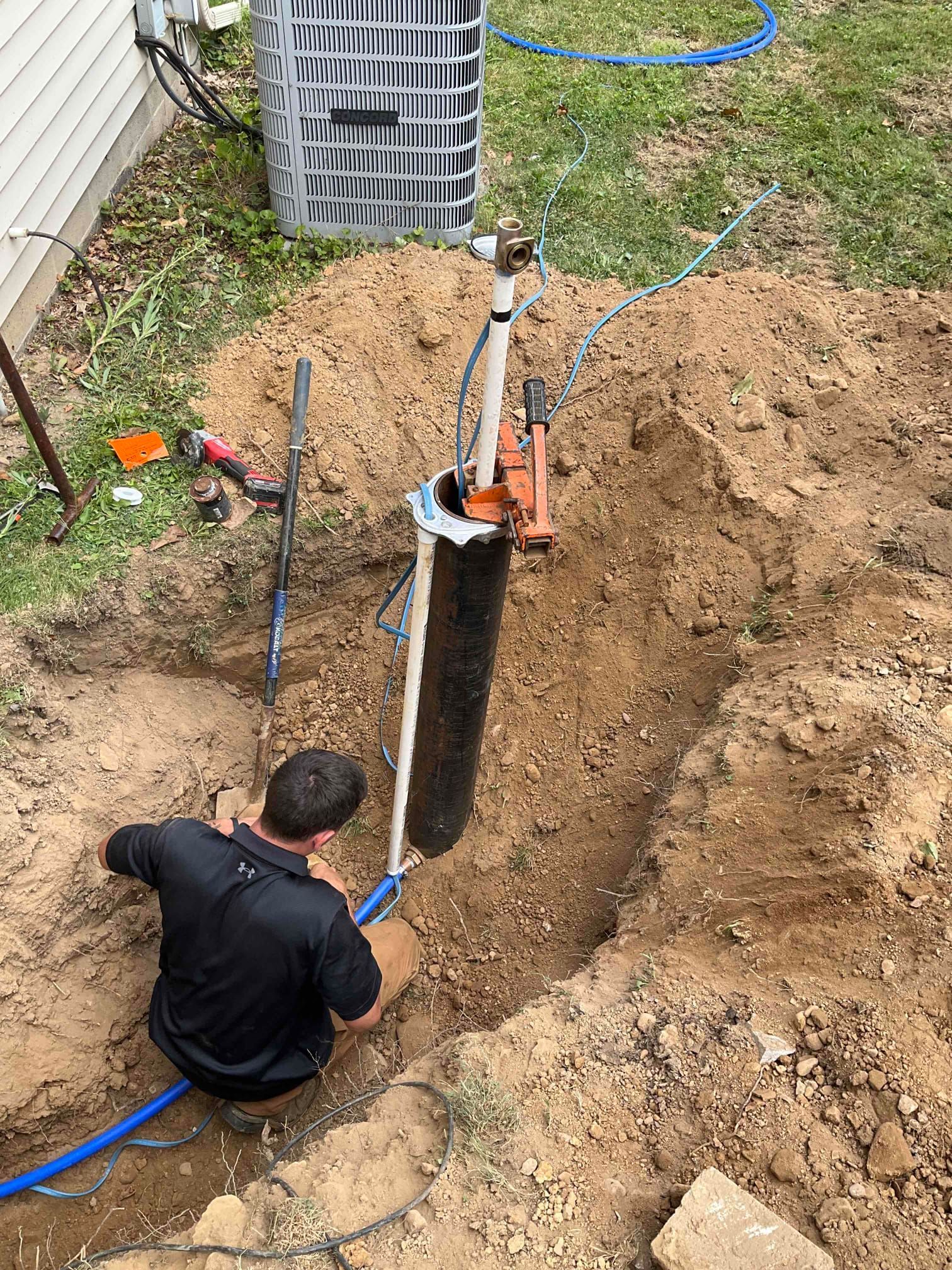A worker kneels in a trench next to a residential water well casing while installing plumbing components.