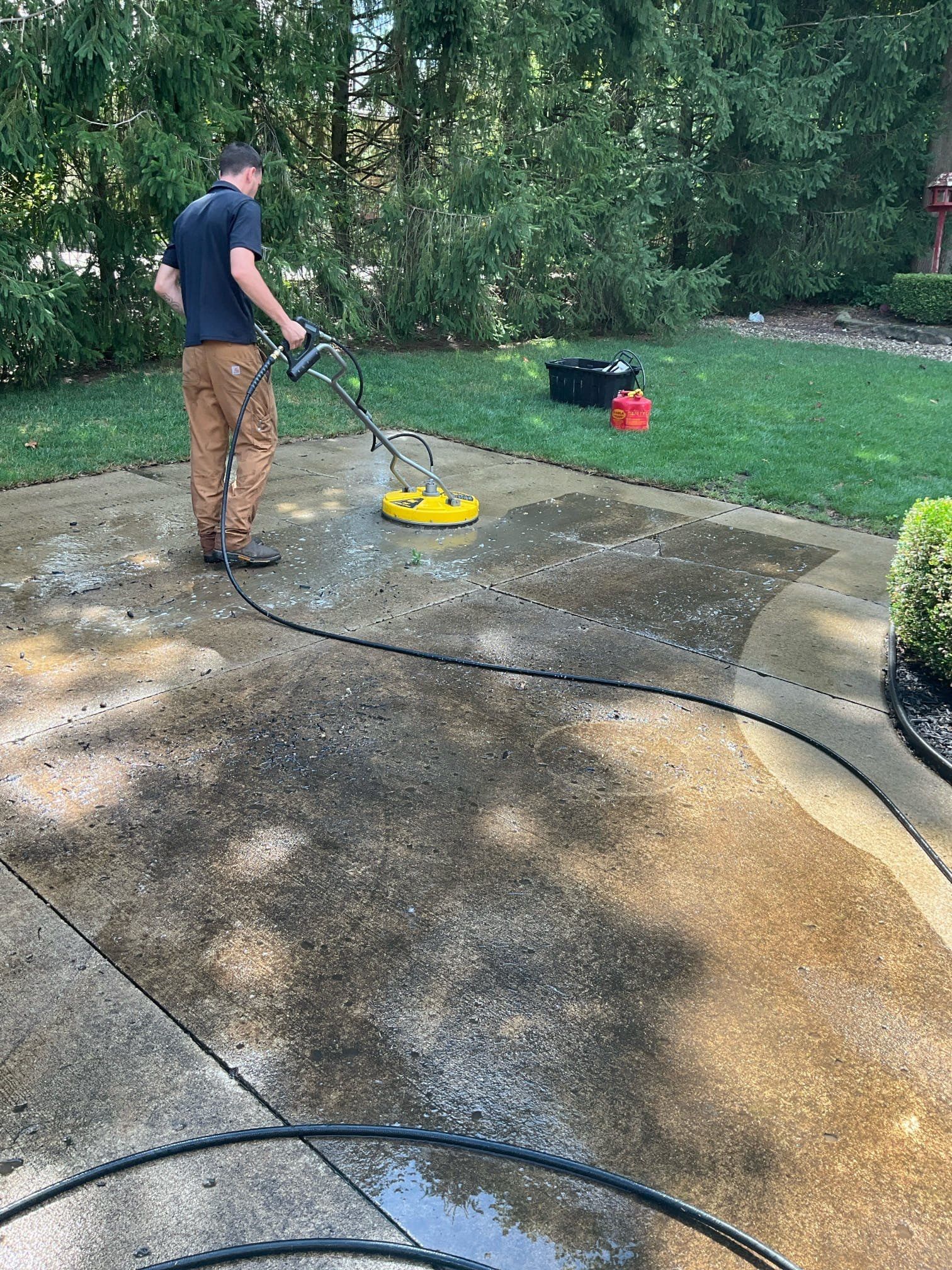 A person using a yellow circular surface cleaner to power wash a concrete driveway in a backyard.