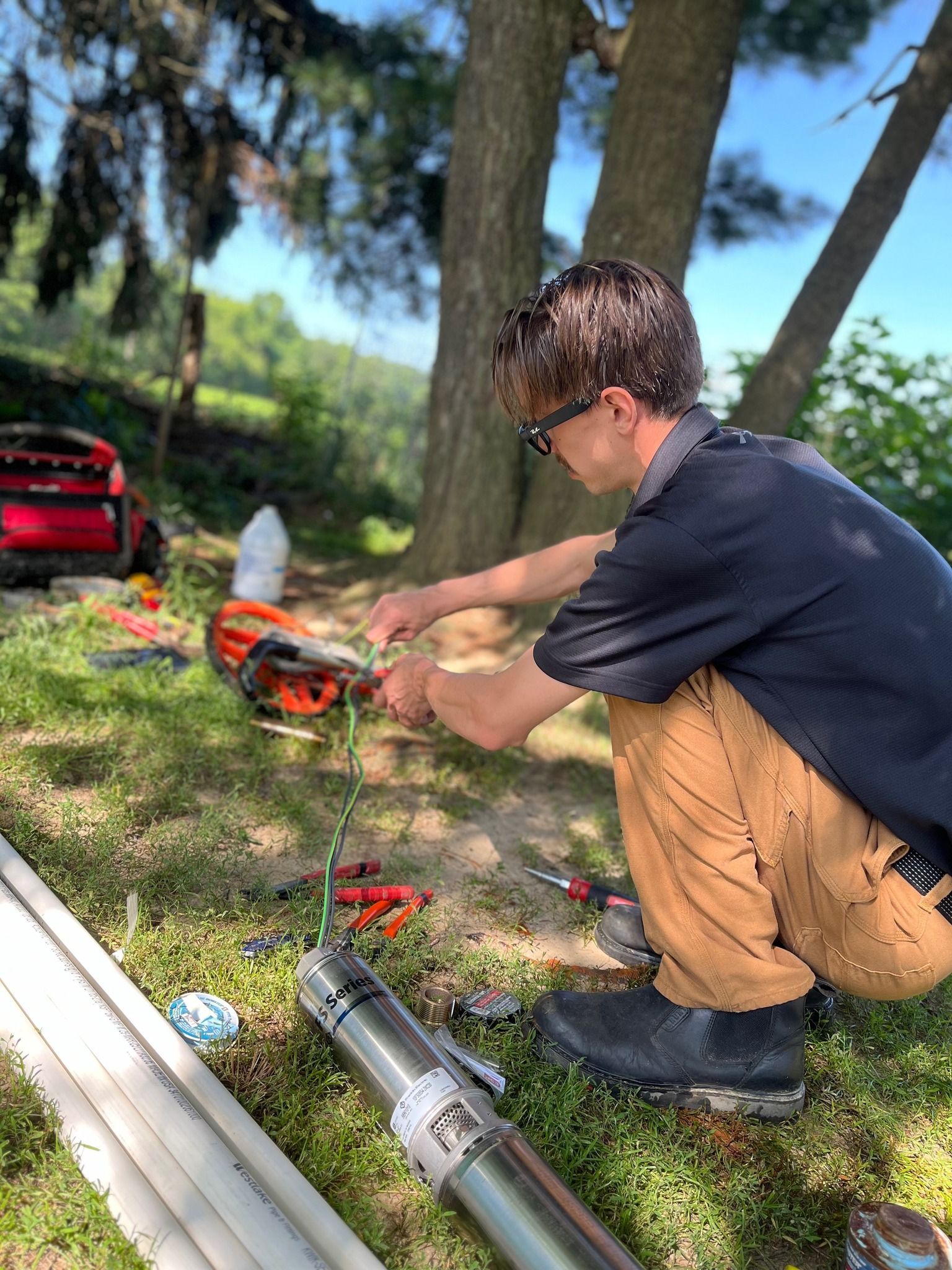 A person in a black shirt and tan pants kneels outdoors, repairing a cylindrical metal well pump on the grass.
