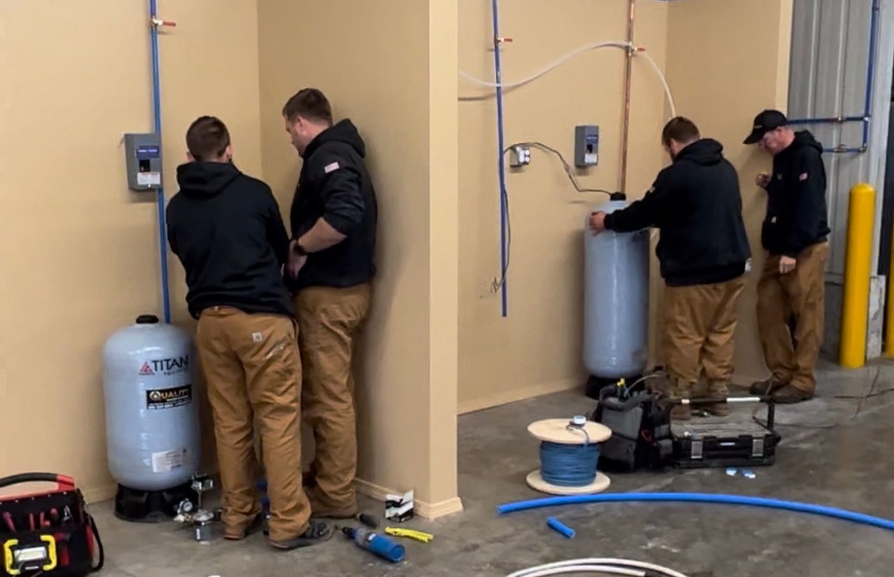 Four people in workwear install water pressure tanks in an unfinished interior space.