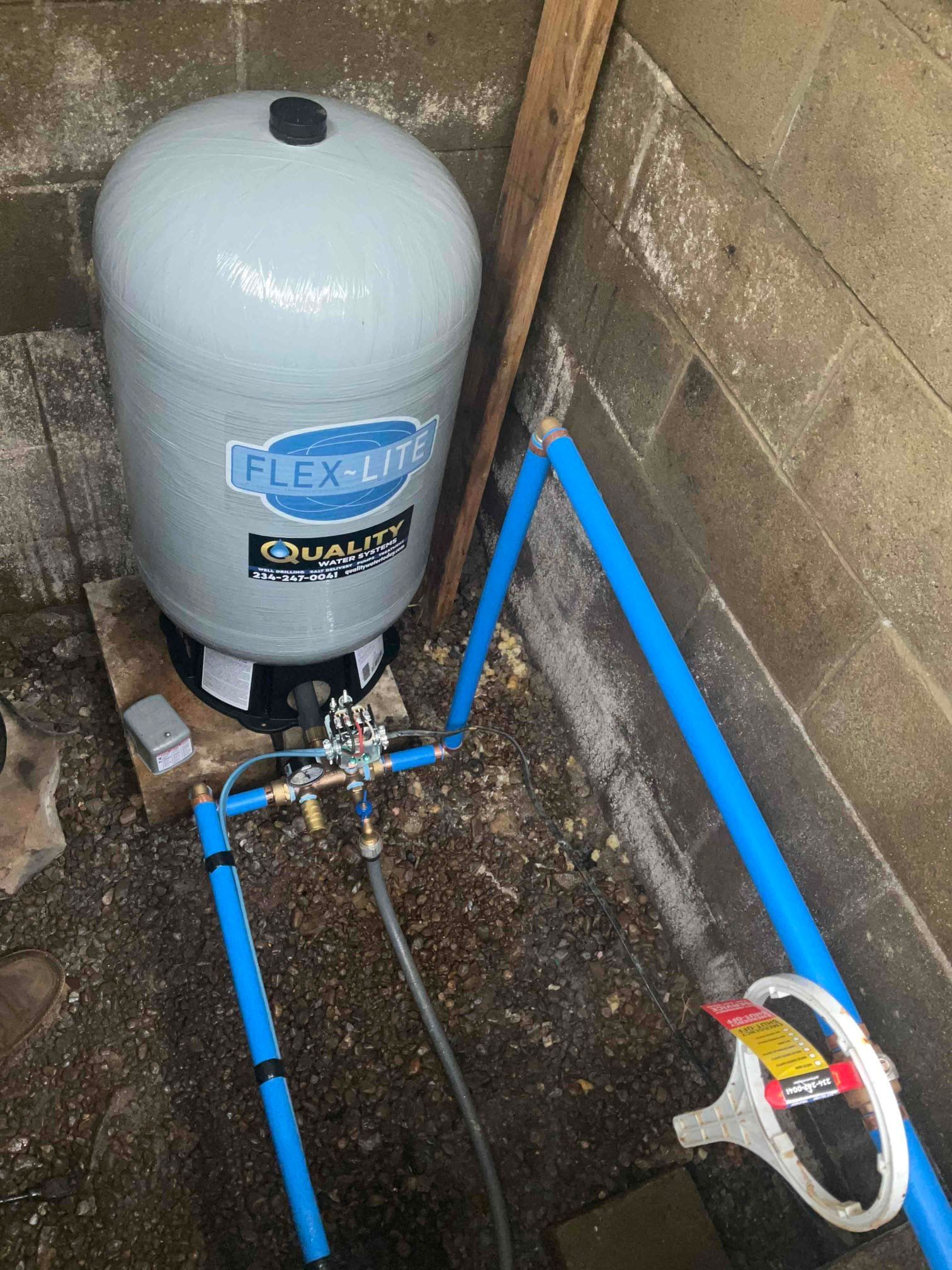 A grey Flex-Lite pressure tank with blue piping and a brass control valve situated in a cinder block corner.