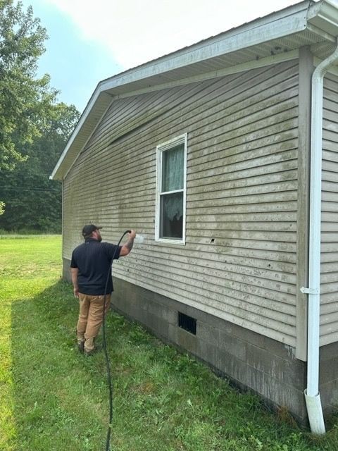 A person in a black shirt and tan pants pressure-washes the dirty, light-colored vinyl siding of a house.