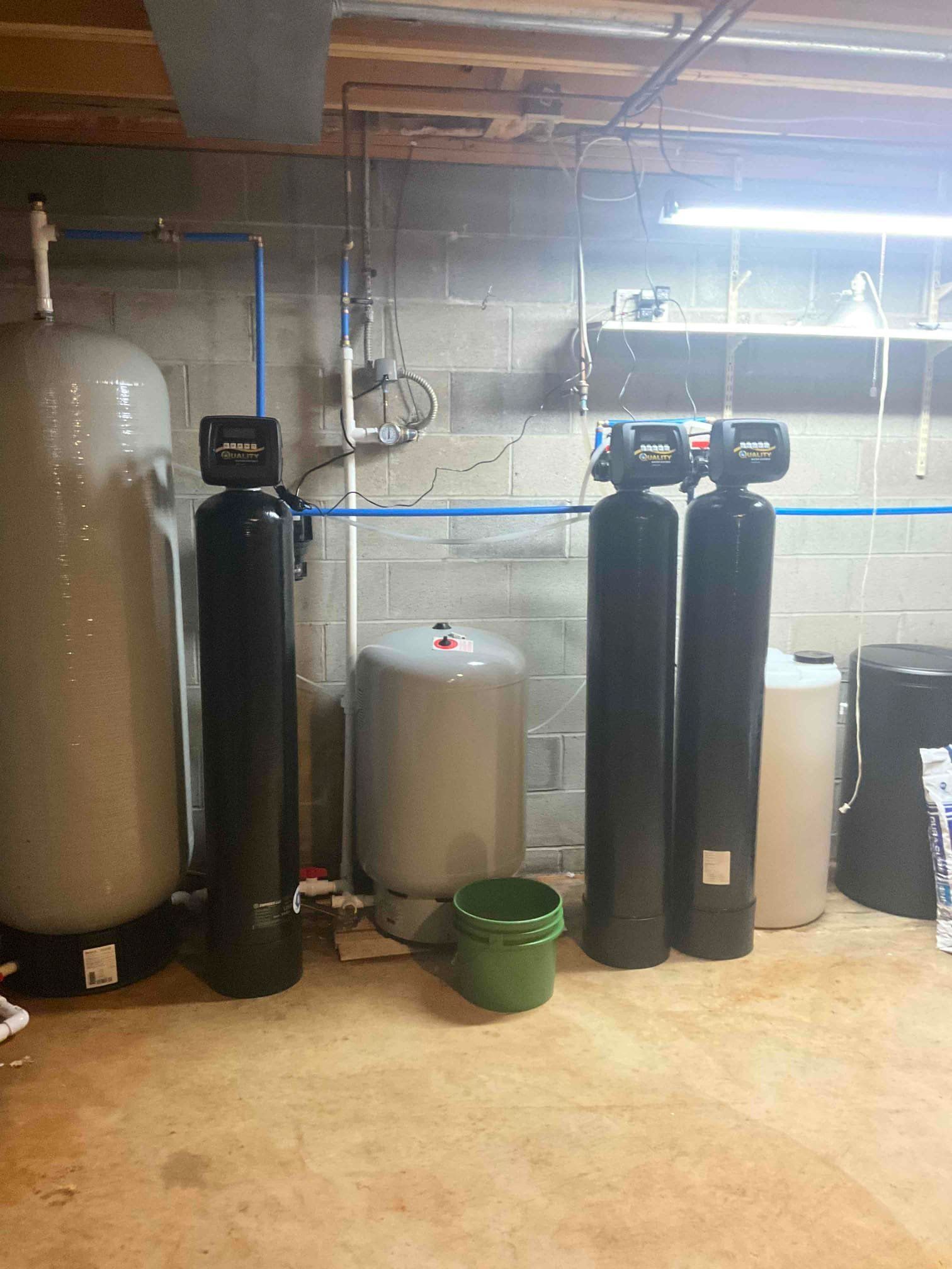 A basement scene with several vertical water filtration tanks, a pressure tank, and a small green bucket on the floor.