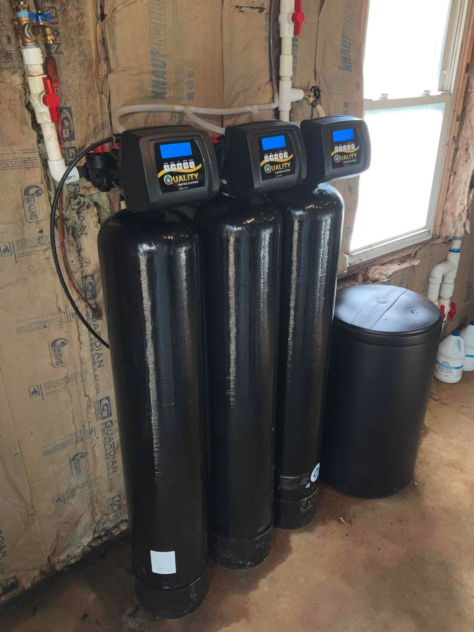 Three black water filtration tanks with digital monitors installed against a wall with insulation and exposed piping.