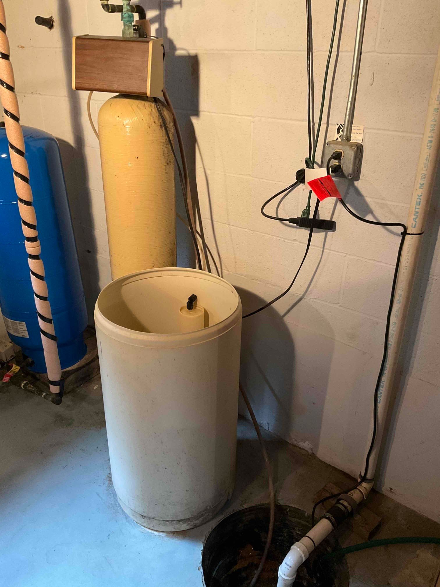 A water softener system with a yellow mineral tank and white brine tank against a concrete block wall in a basement.