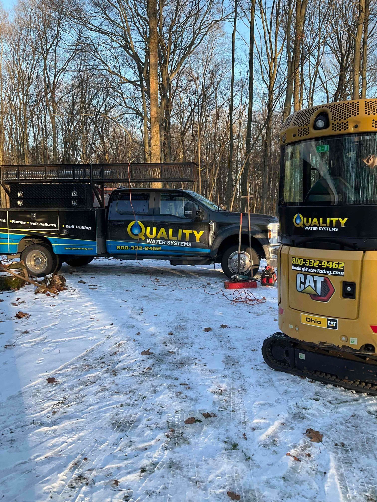 A black utility truck and a yellow CAT mini excavator parked on snow-covered ground in front of a forest.