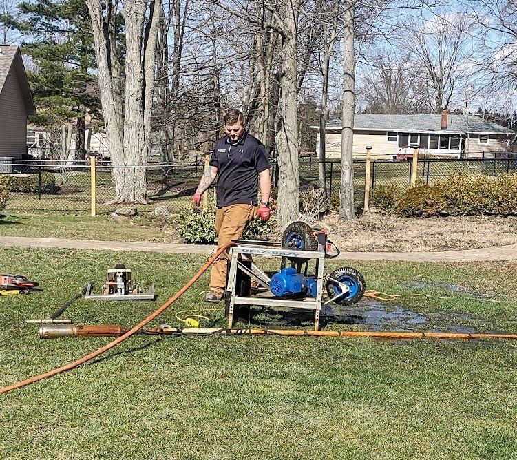 A person in a black shirt and tan pants operates a pipe-cleaning machine in a grassy yard near a suburban house.
