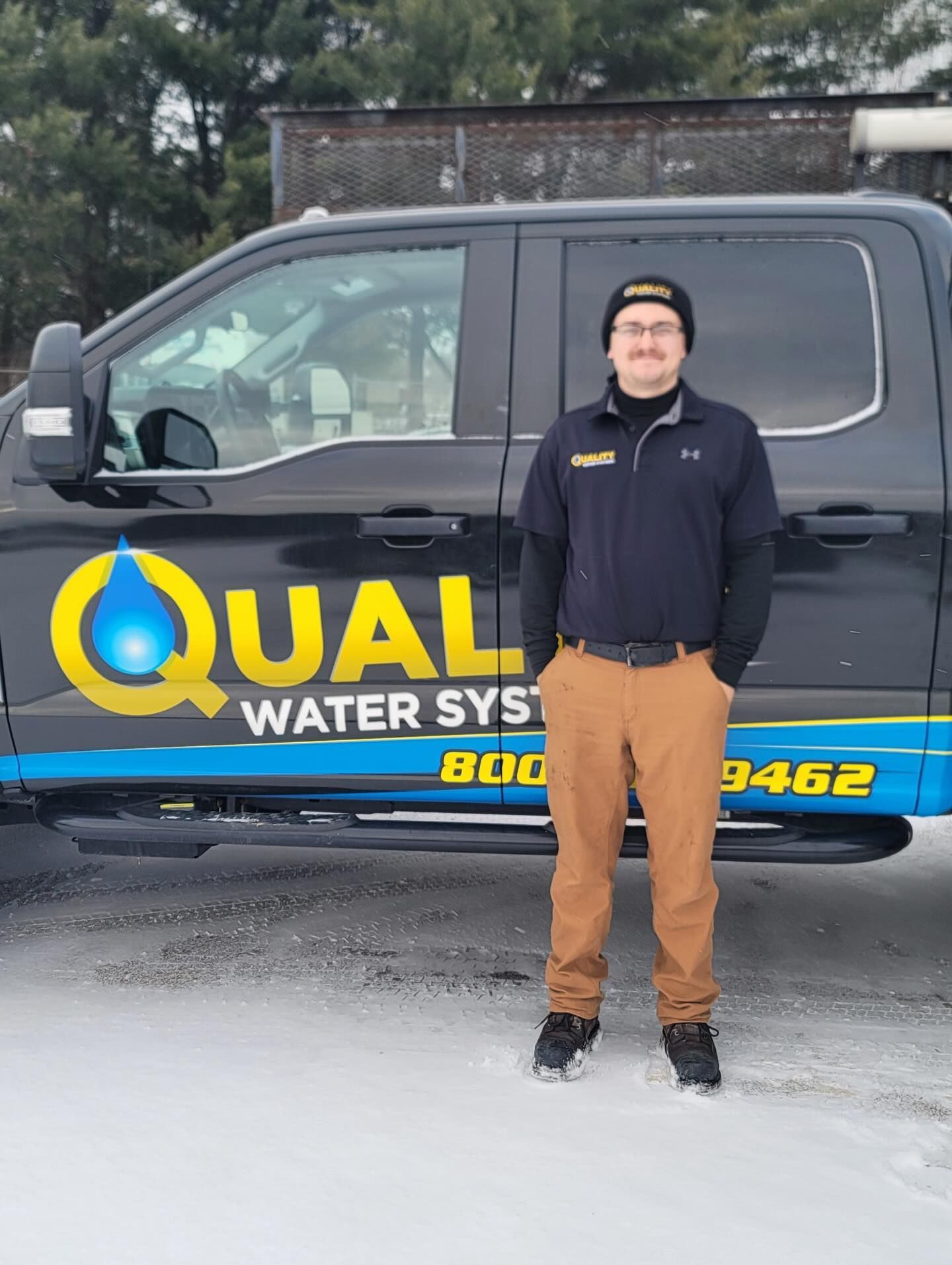 A person wearing a beanie and uniform stands in the snow in front of a black company truck labeled Quality Water Systems.