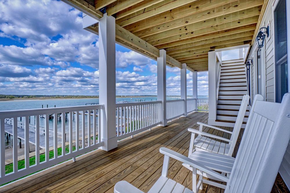 There are two rocking chairs on the deck of a house with a view of the ocean.