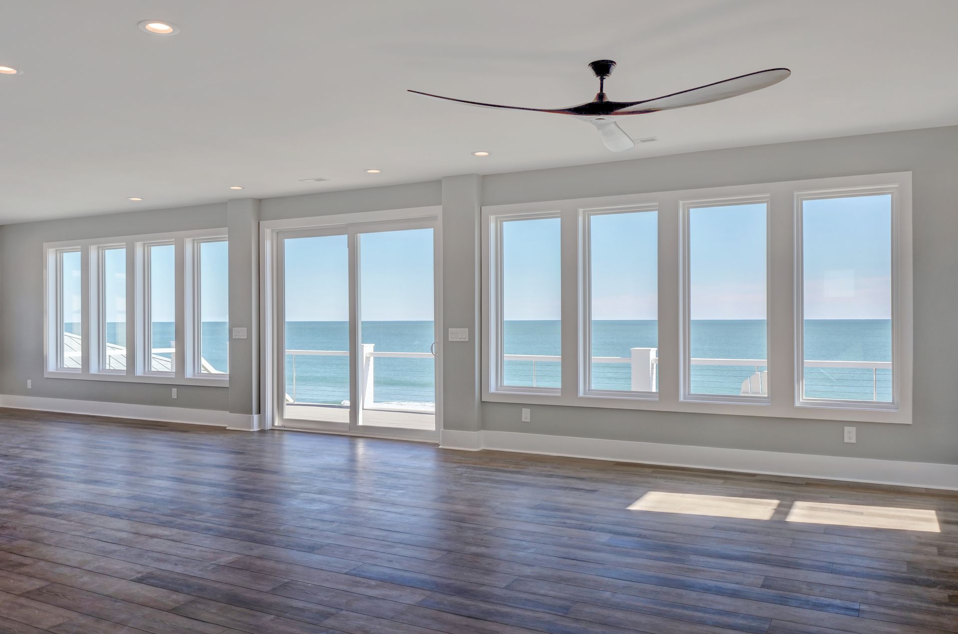 An empty living room with a ceiling fan and lots of windows overlooking the ocean.