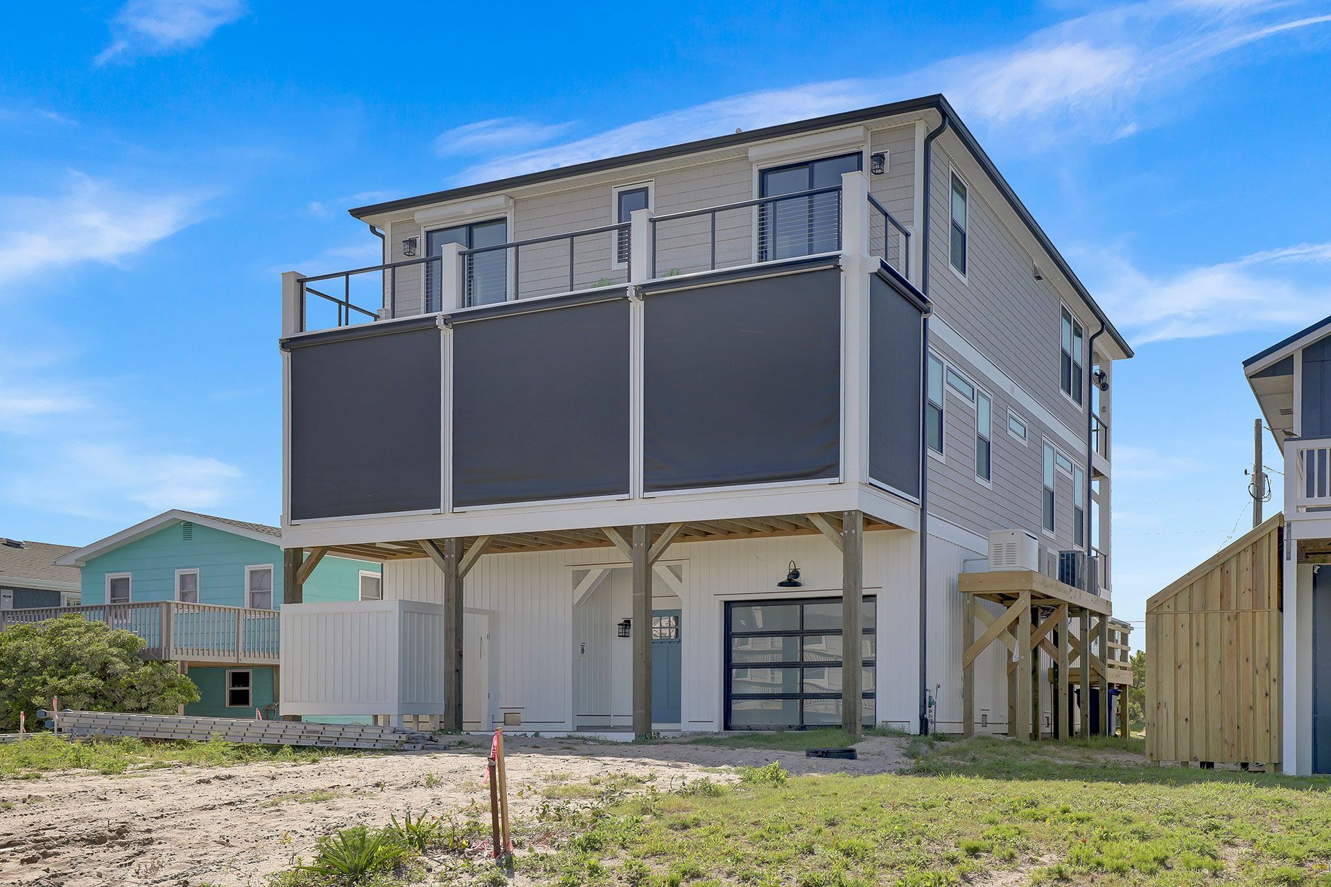 Three-story beach house with gray siding, glass garage door, and outdoor decks, on a sandy lot under a blue sky.