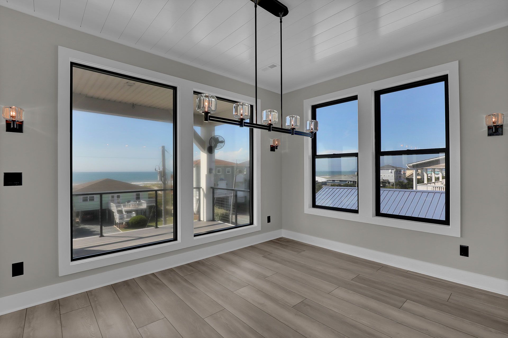 Empty room with large windows overlooking the ocean. Gray walls, wood floors, and a modern chandelier.