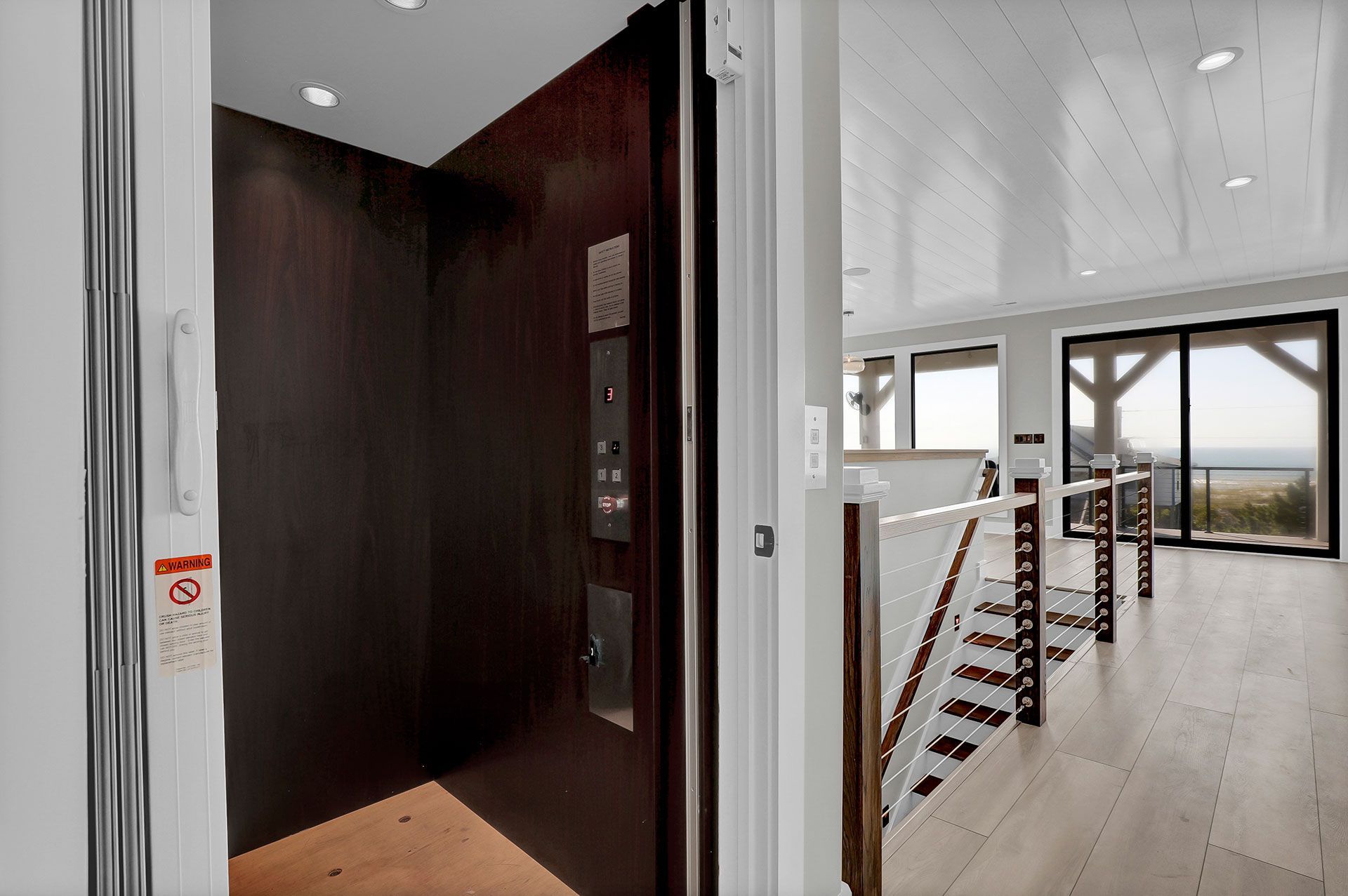 Elevator with dark wood paneling, next to stairs, and light-filled living area with ocean view.