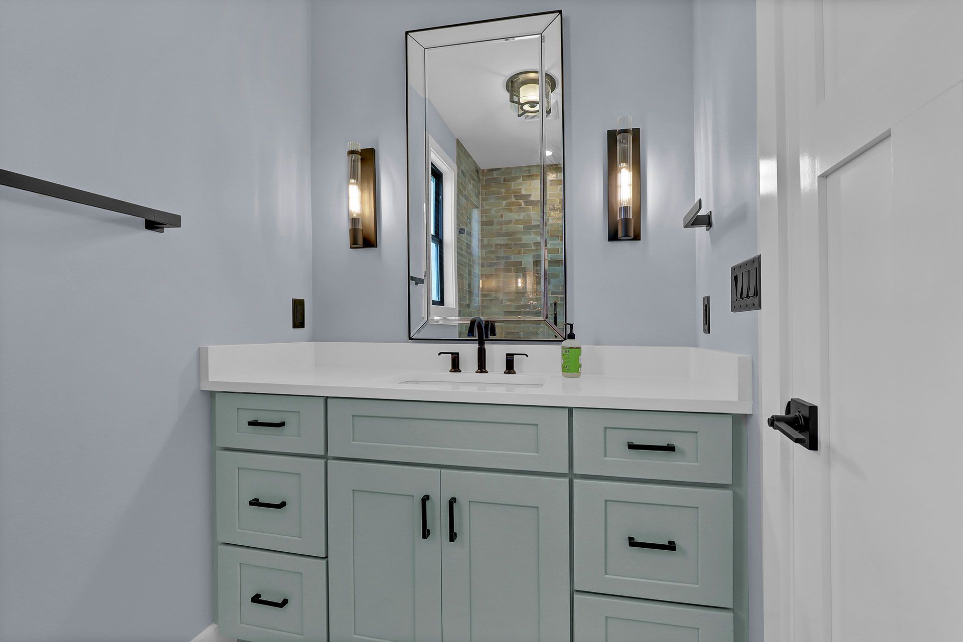 Bathroom with a light blue wall, a green vanity with a white countertop, a mirror, and sconces.