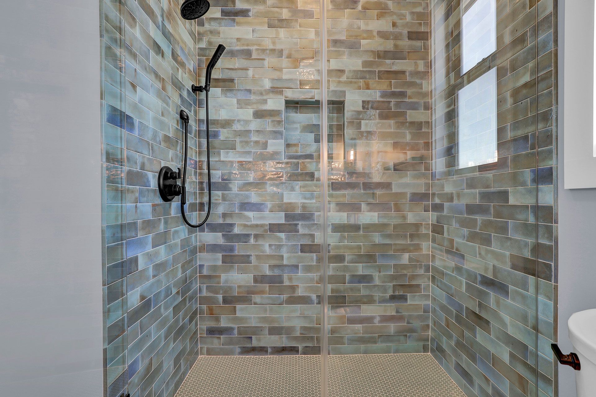 Shower with blue-gray brick tile, black fixtures, and glass door. Small recessed shelf and pebble floor.