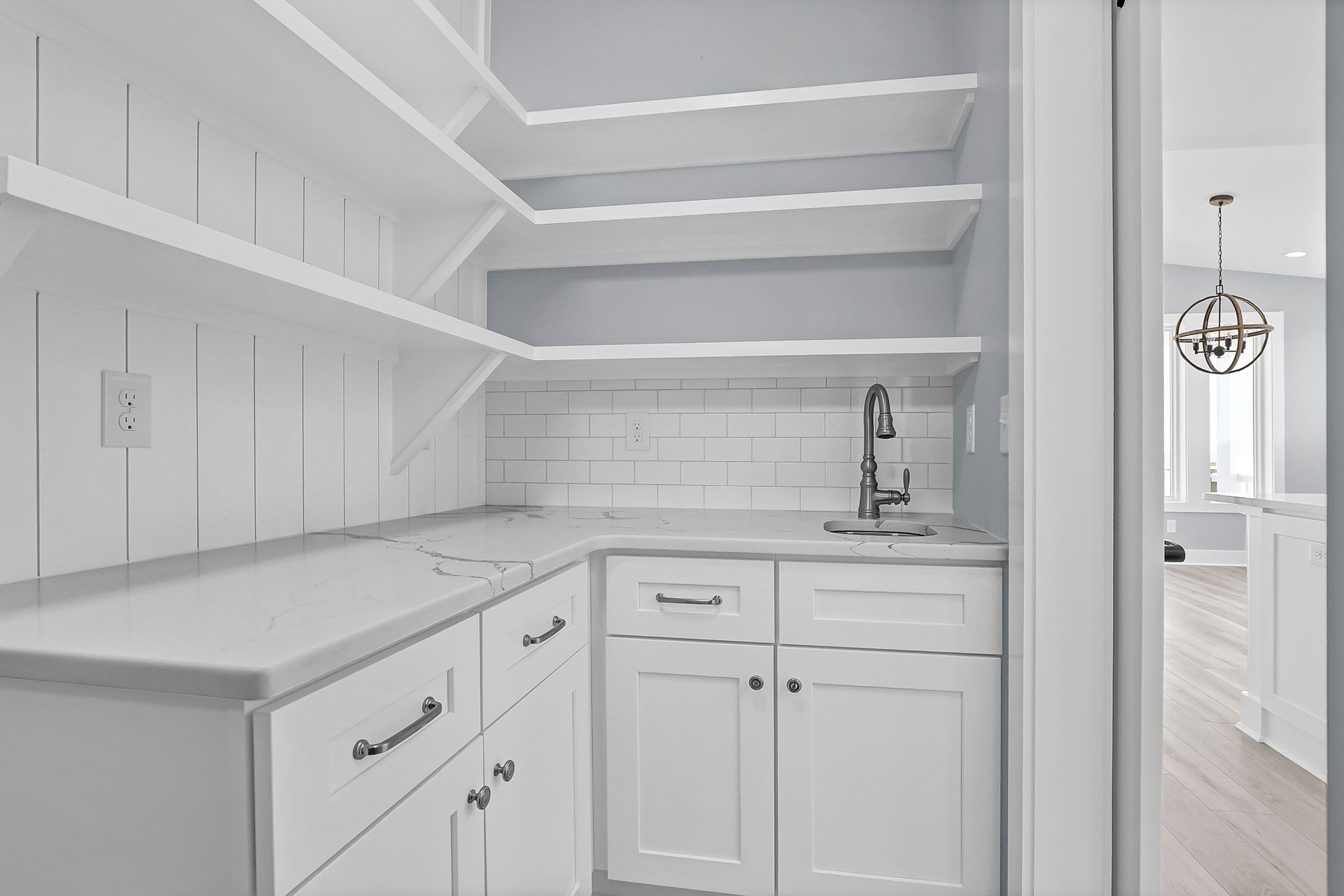 White pantry with shelves, cabinets, and sink, opening to a brighter room.