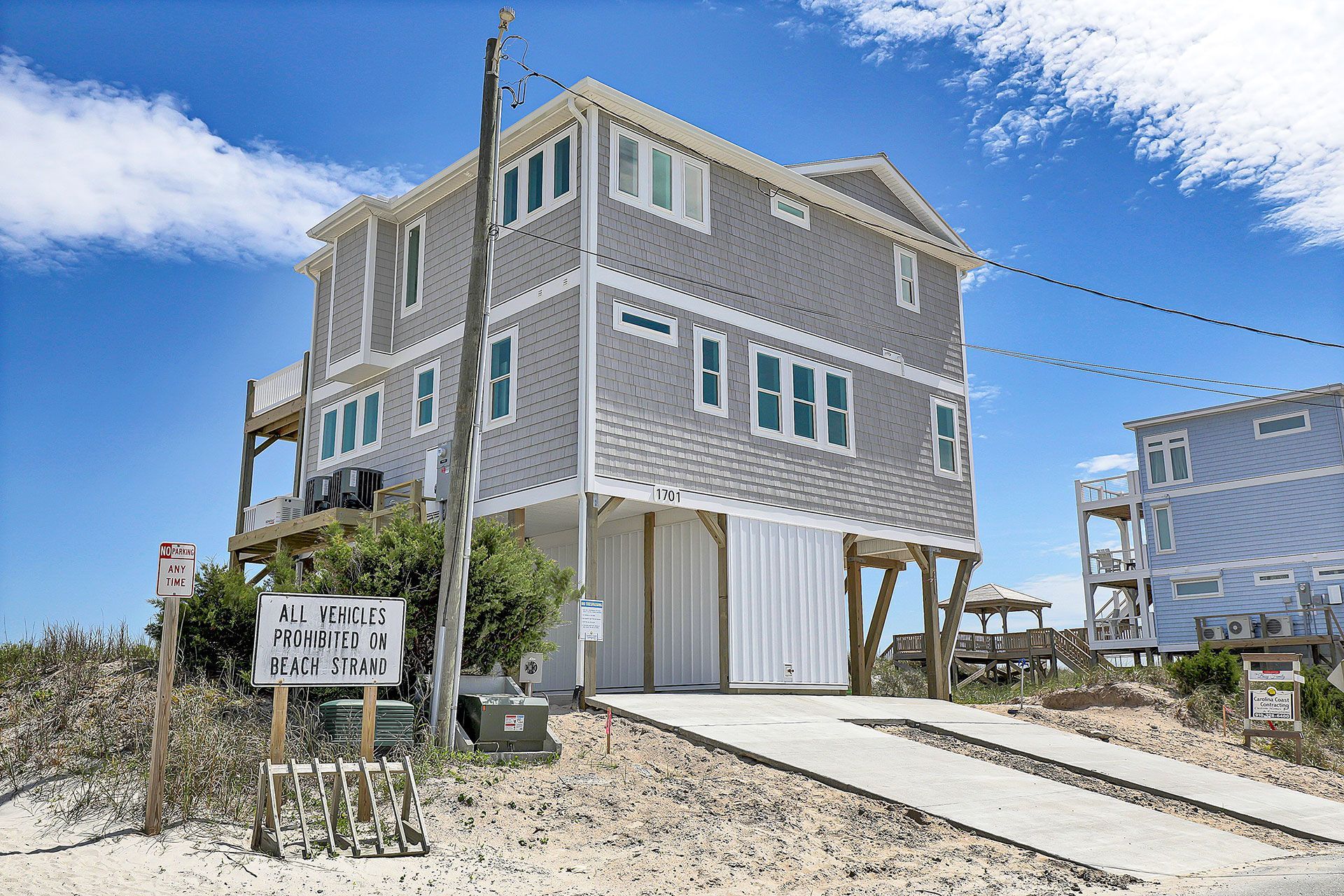 A three-story gray beach house on stilts, with a driveway, mailbox, and blue sky.