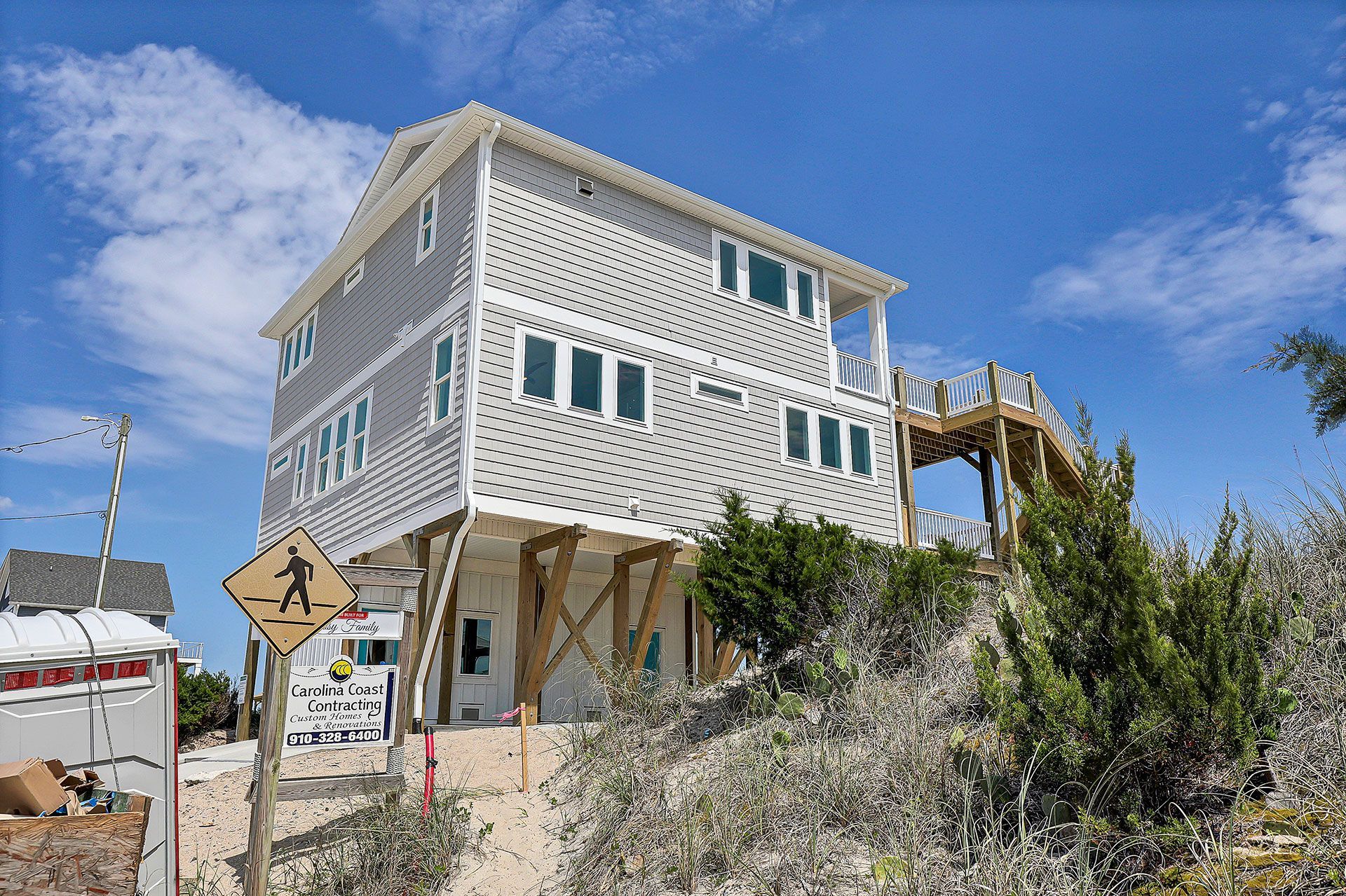Three-story gray beach house on stilts with boardwalk access, set against a blue sky.