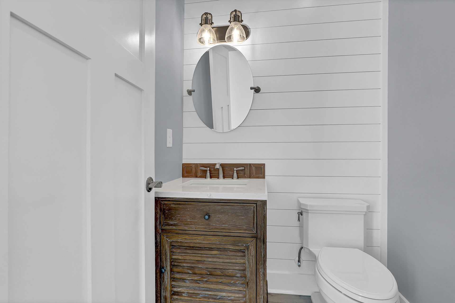 A small bathroom with a rustic vanity, toilet, oval mirror, and shiplap wall.