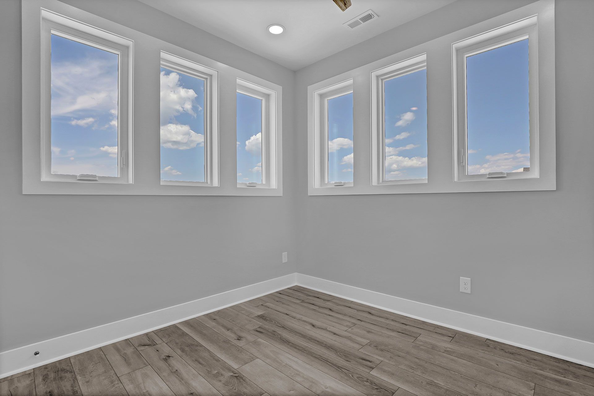 Empty room with multiple windows, light gray walls, and wood-look flooring; blue sky visible through windows.