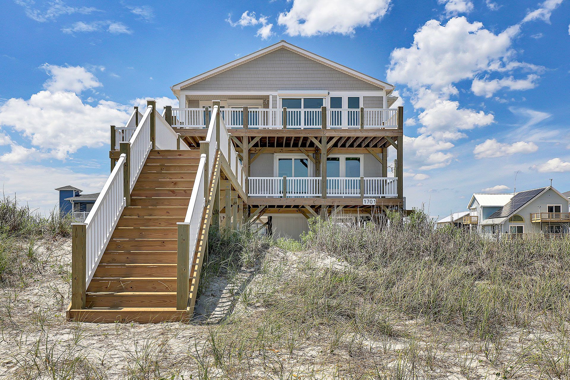 Two-story beach house with wooden stairs and balconies, set amidst sand and dune plants under a blue sky.