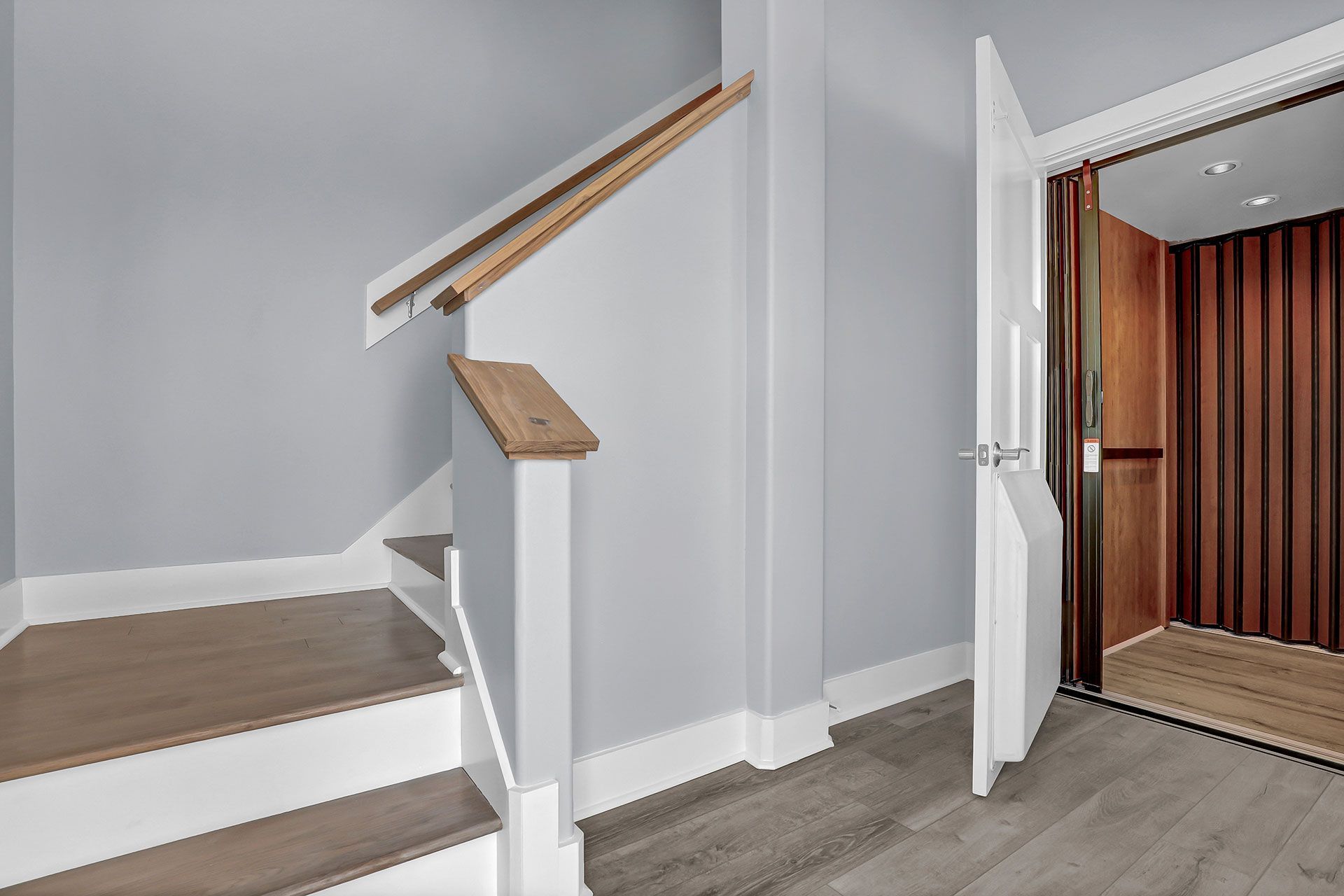Staircase with wooden steps and railing next to an open elevator in a hallway with gray walls.