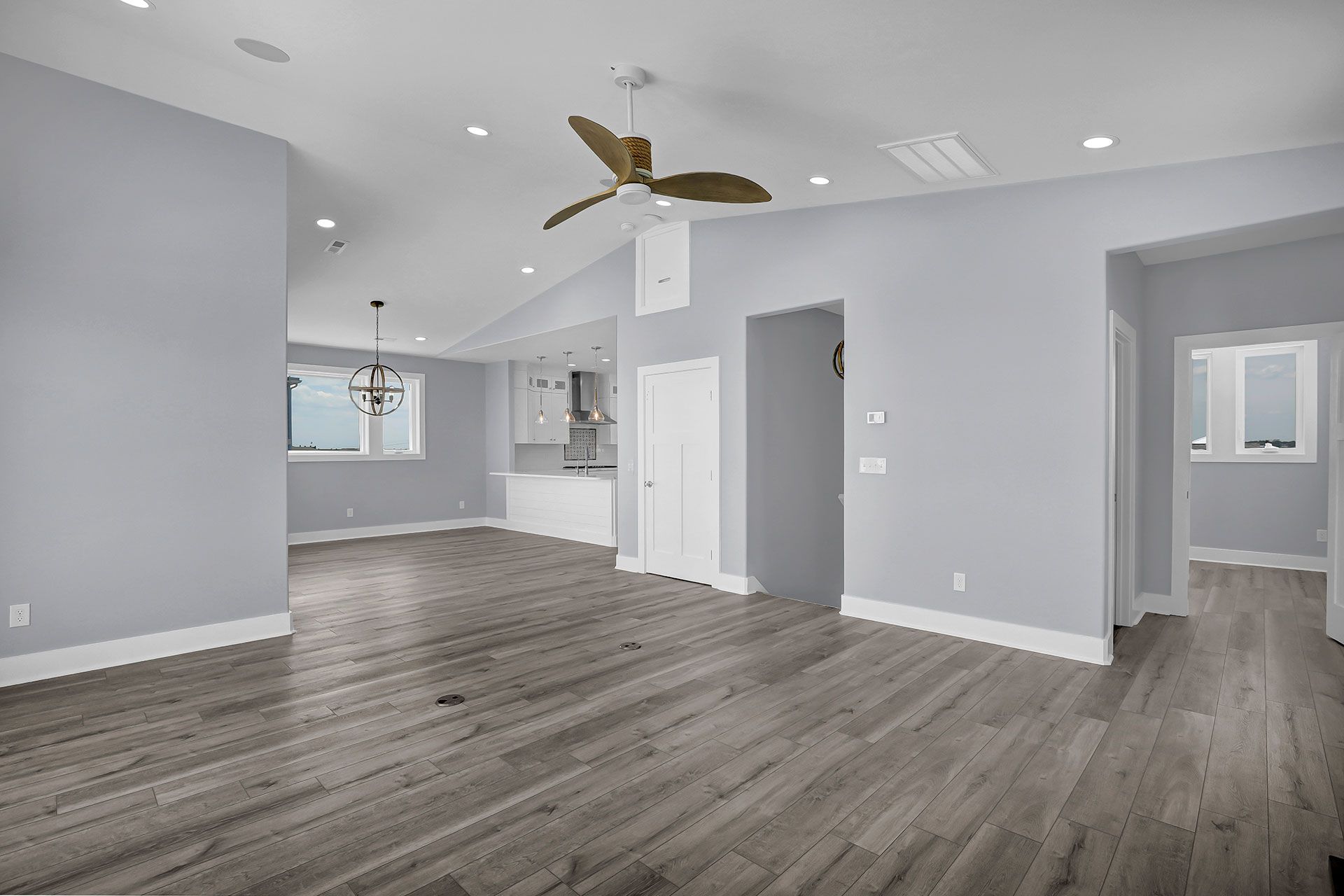Empty, light-filled room with gray wood floors, pale blue walls, and a ceiling fan; doors lead to other rooms.