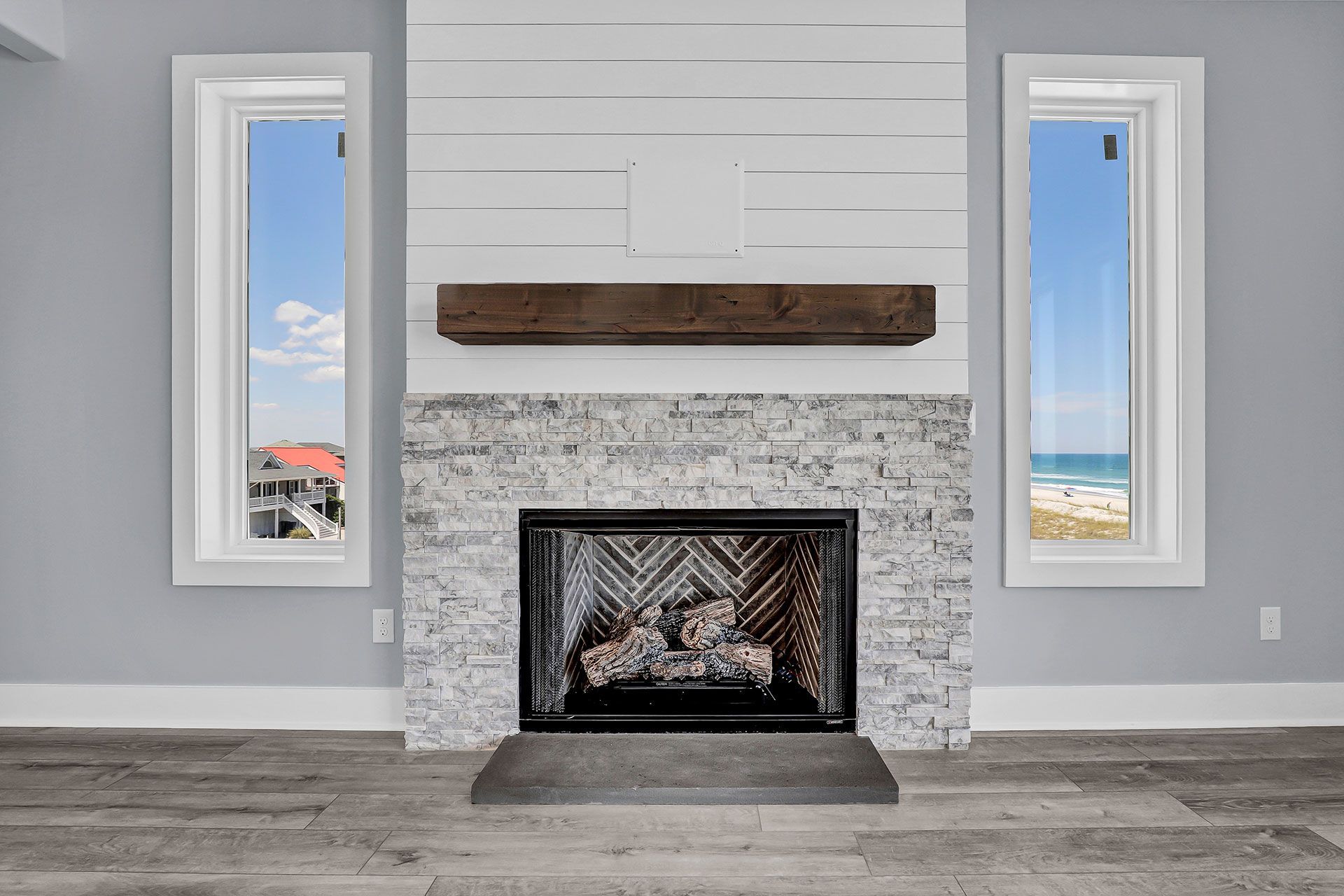 Fireplace with stone surround, wooden mantel, flanked by windows with ocean views.