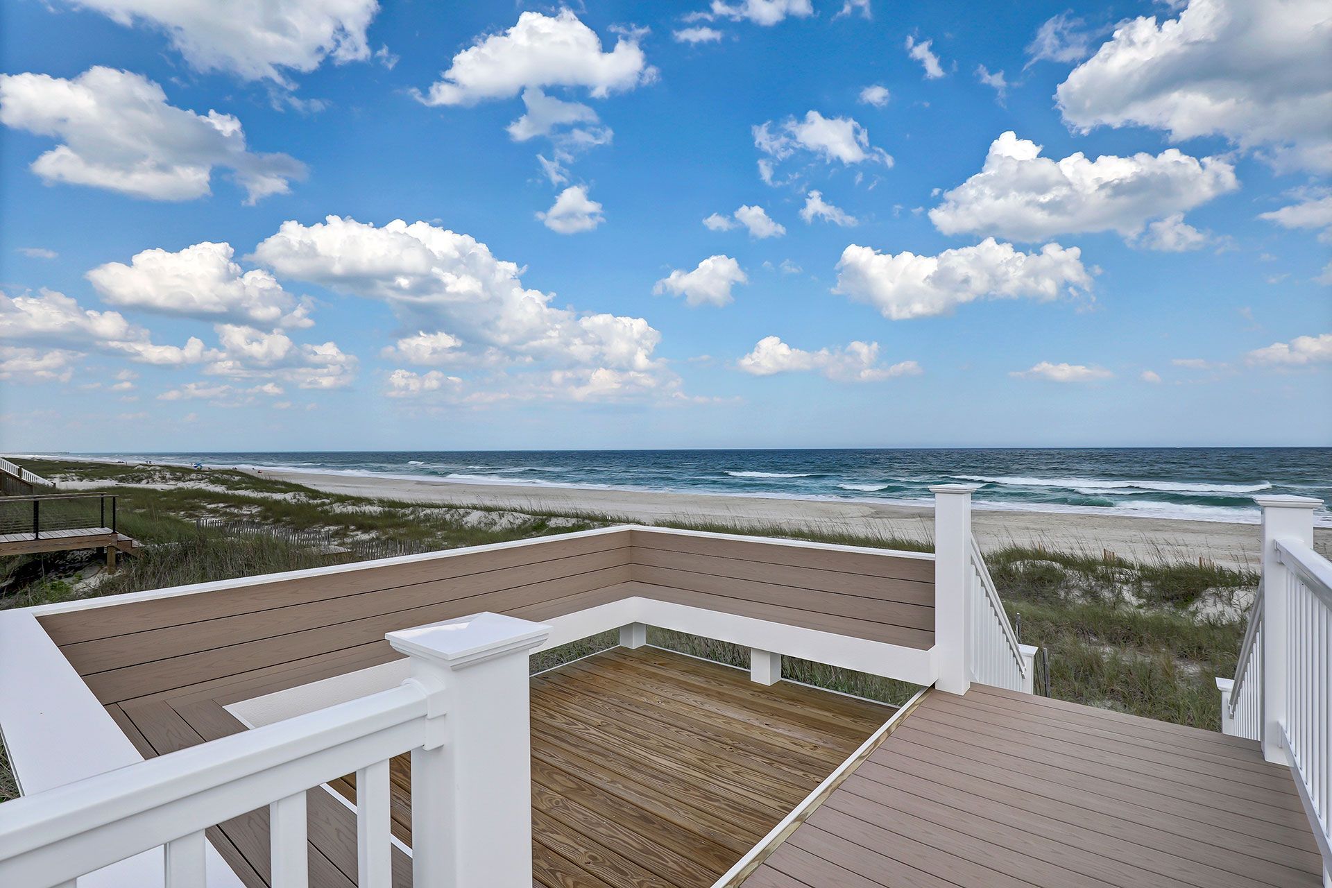 A beach view from a deck, with blue sky, ocean, and sand dunes.
