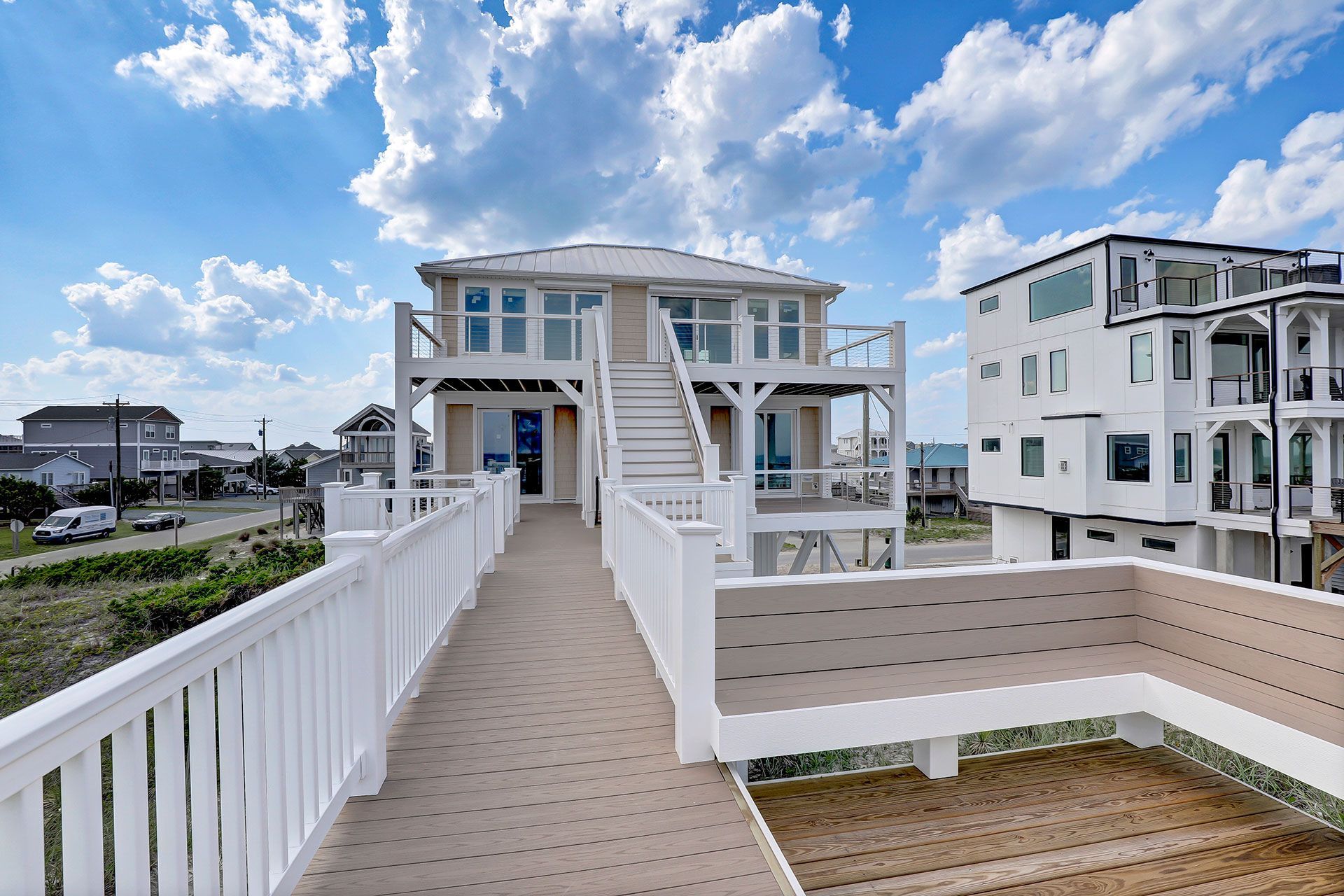 Exterior view of a beach house with a deck and stairs, against a blue sky with clouds.