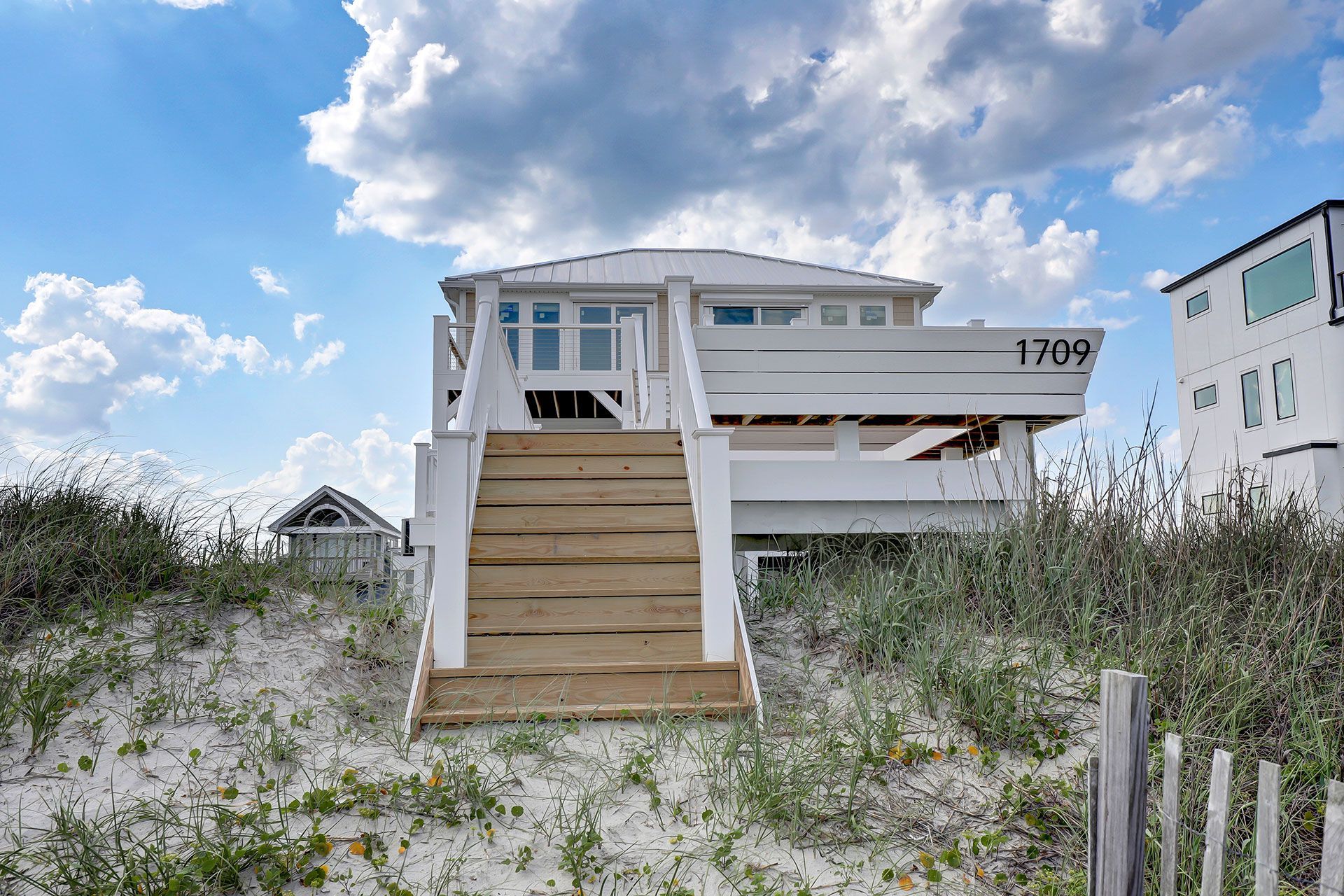 White beach house with wooden steps leading up, set in sand dunes.