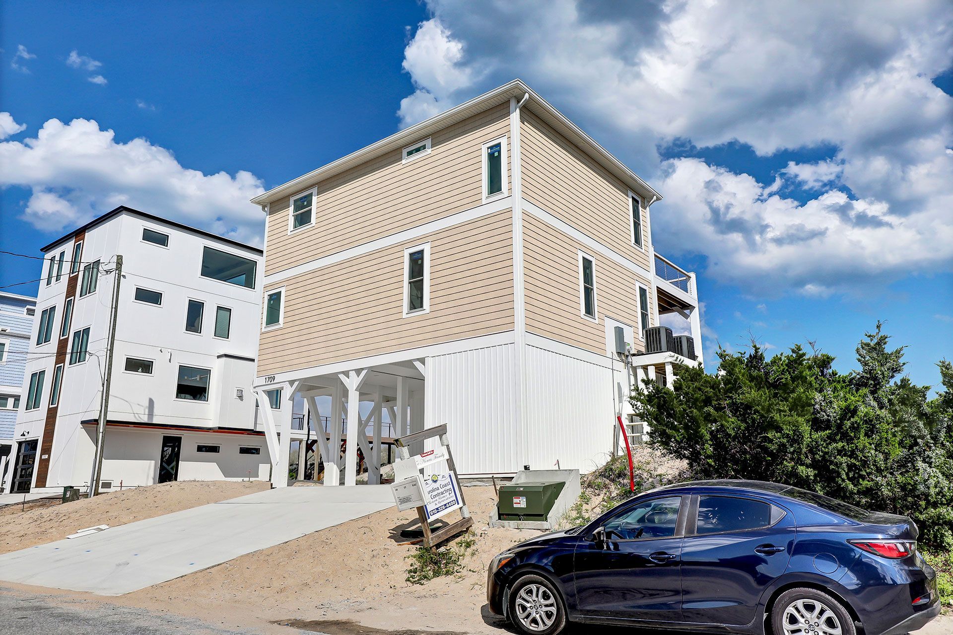 Blue car parked in front of a three-story coastal home with tan siding and a blue sky.
