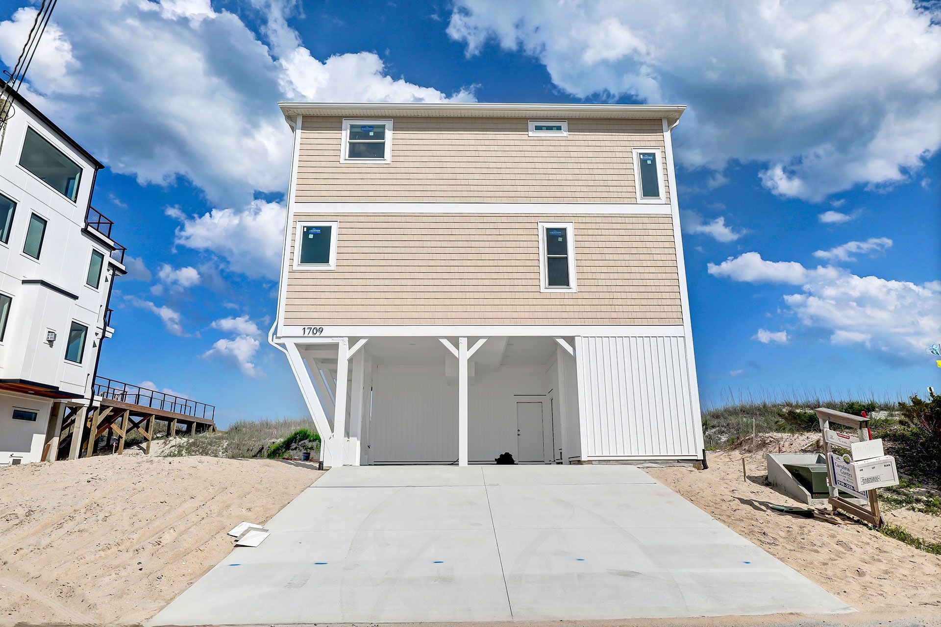 A two-story beach house with a concrete driveway. Beige siding, white accents, and a blue sky.