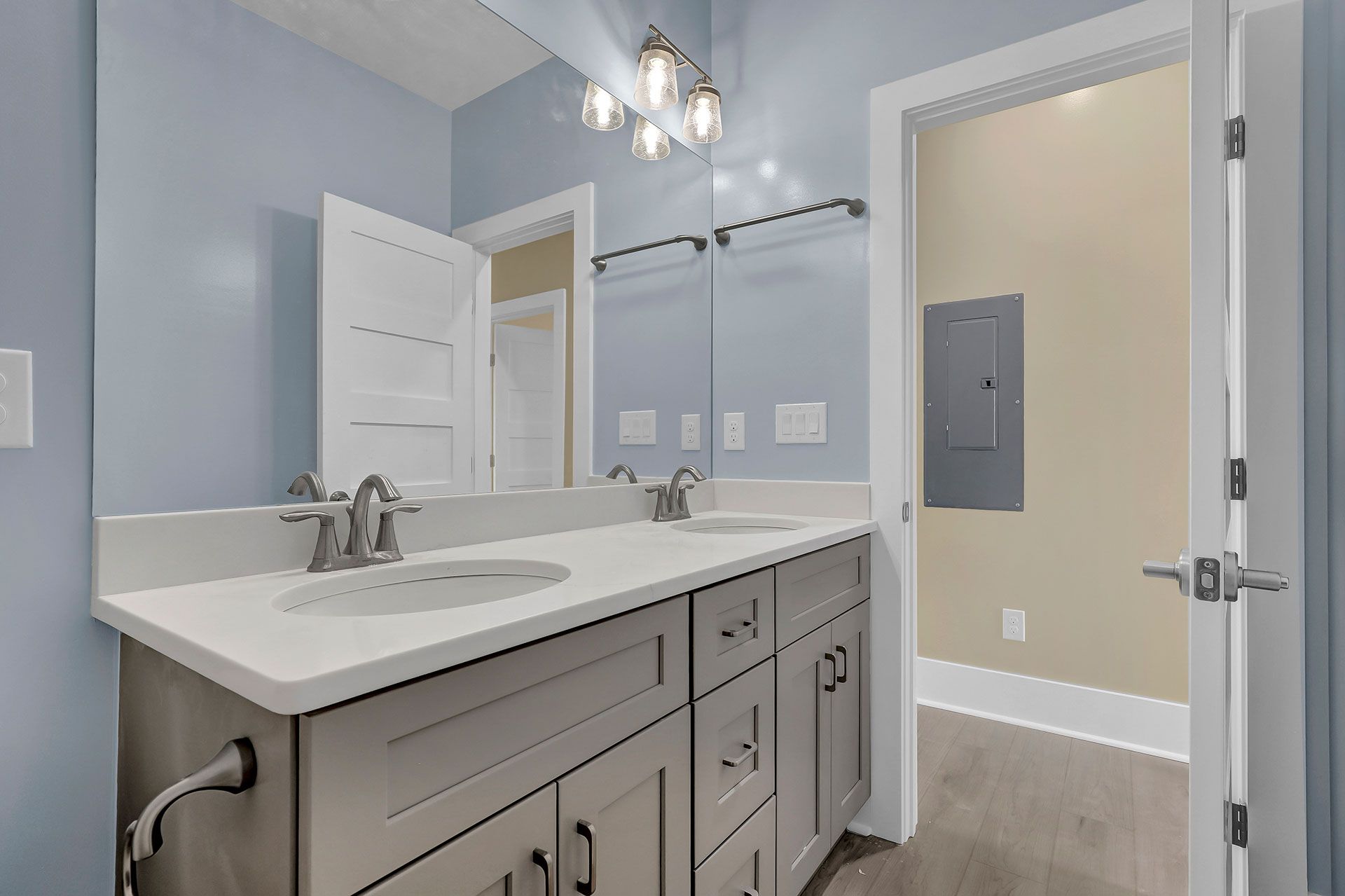 Bathroom with light blue walls, double sinks, and open door to another room.