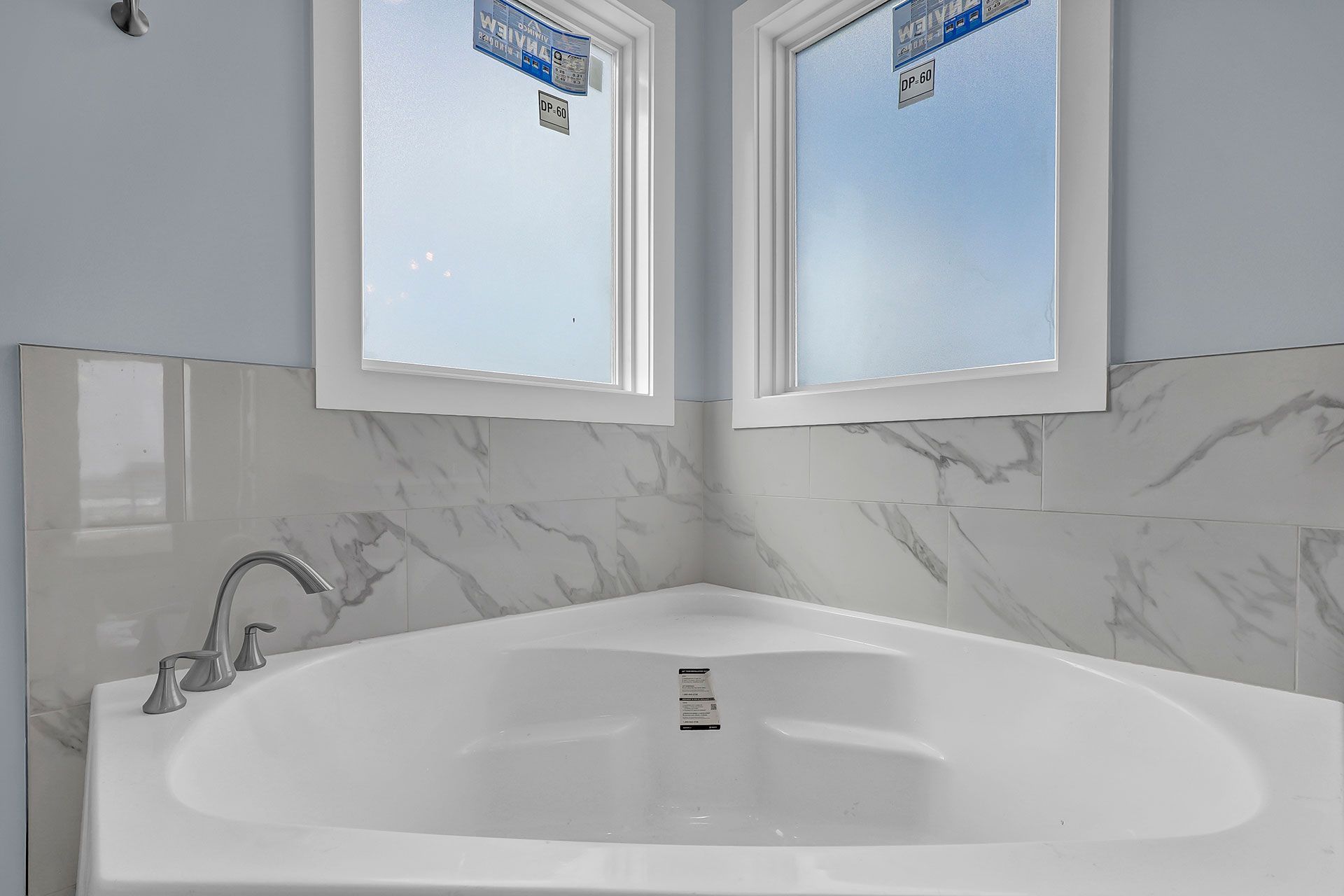 A white bathtub in a corner with marble tiles, two windows, and light blue walls.