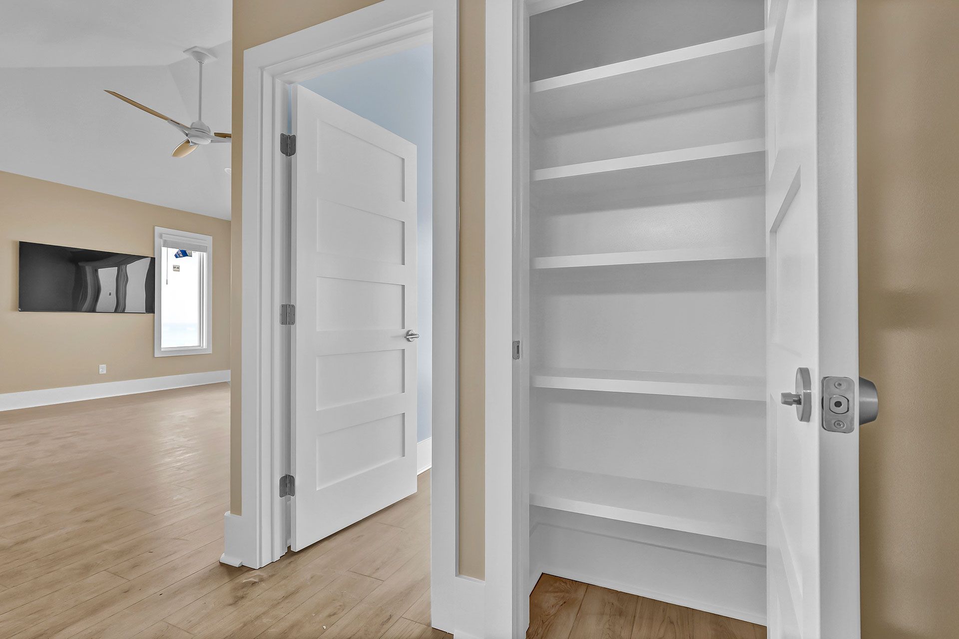 Open pantry with empty white shelves next to a partially open white door, leading into a room with a tv.