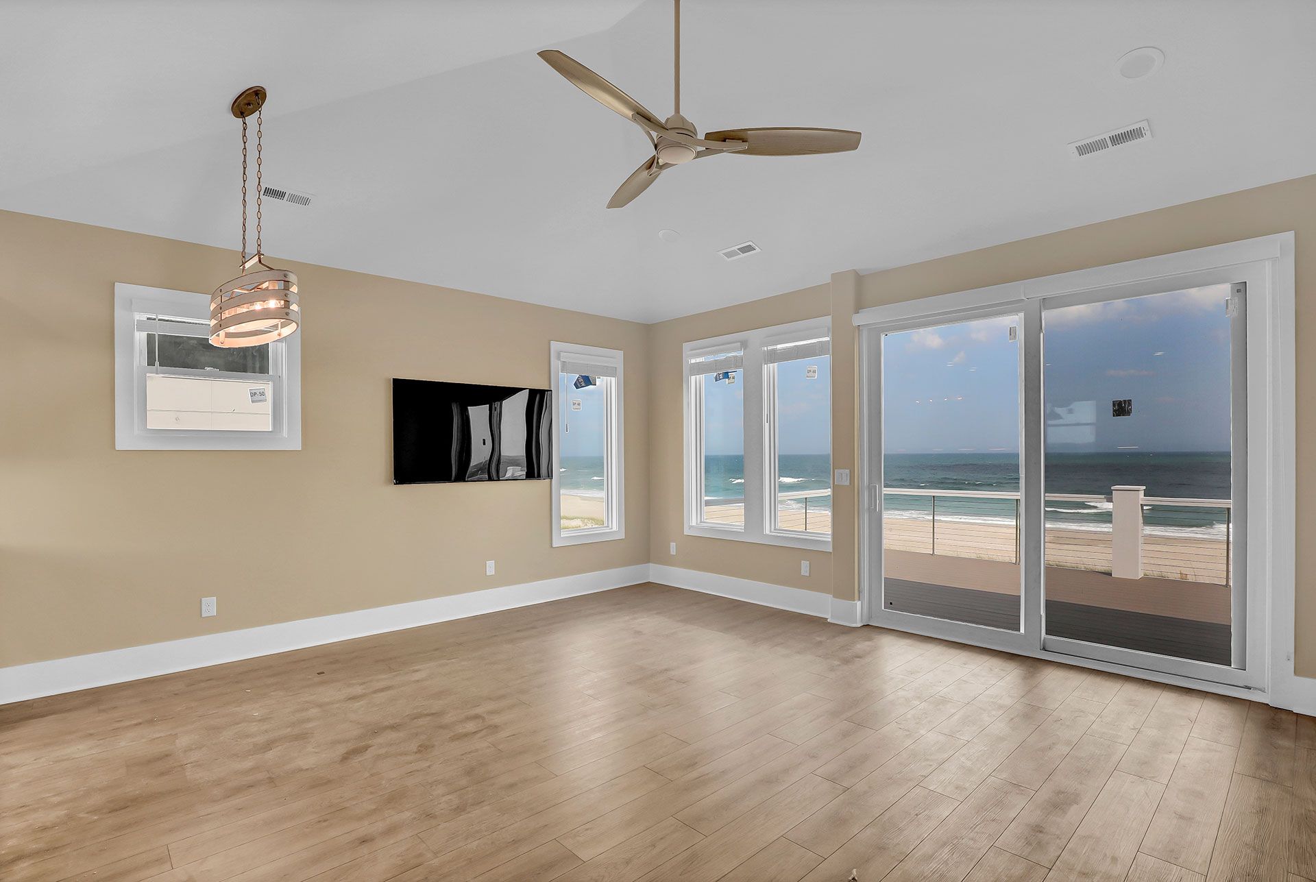 Empty room with ocean view through sliding glass doors. Light wood floors, beige walls, ceiling fan.