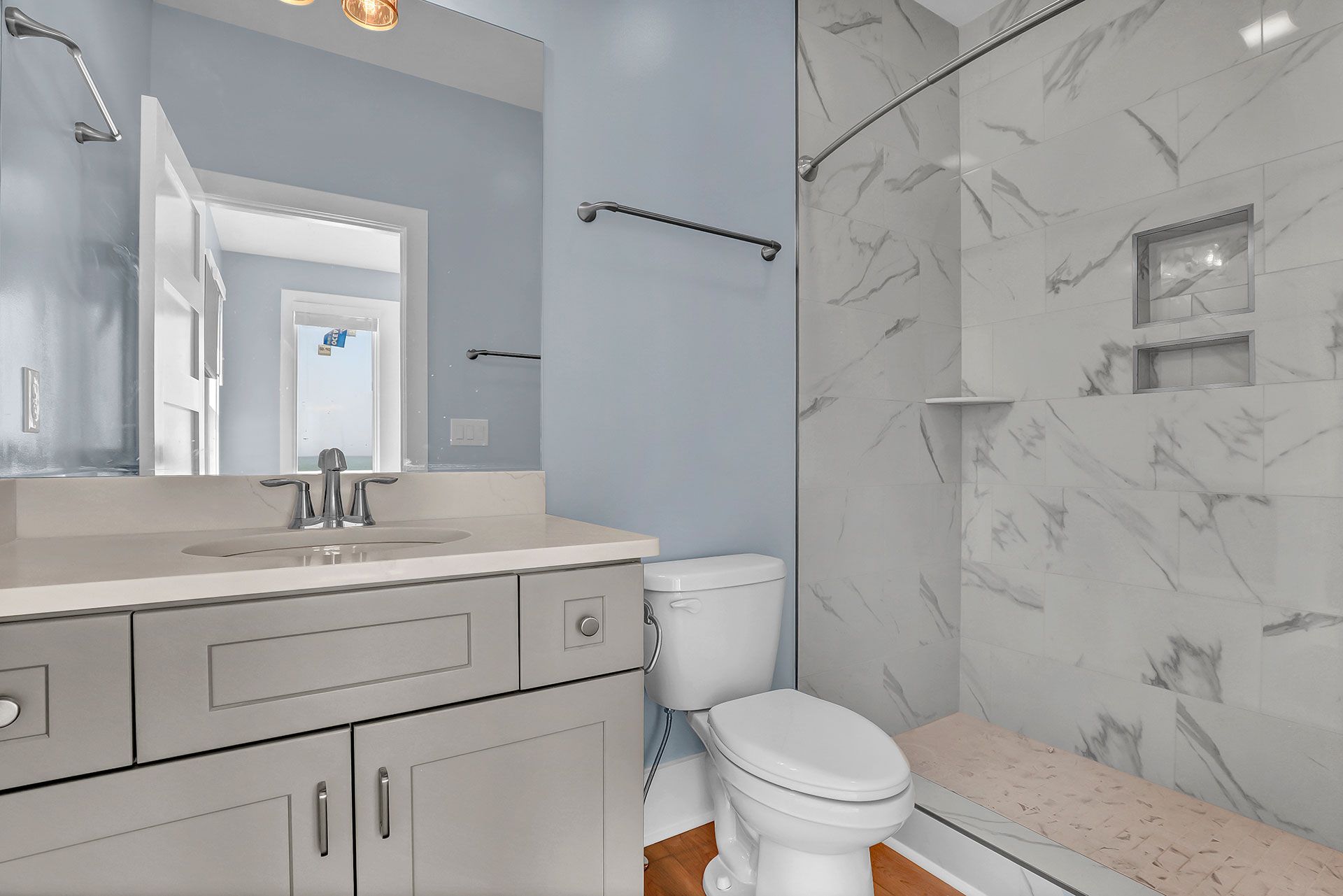 A modern bathroom with a light blue wall, white vanity, marble shower, and toilet.
