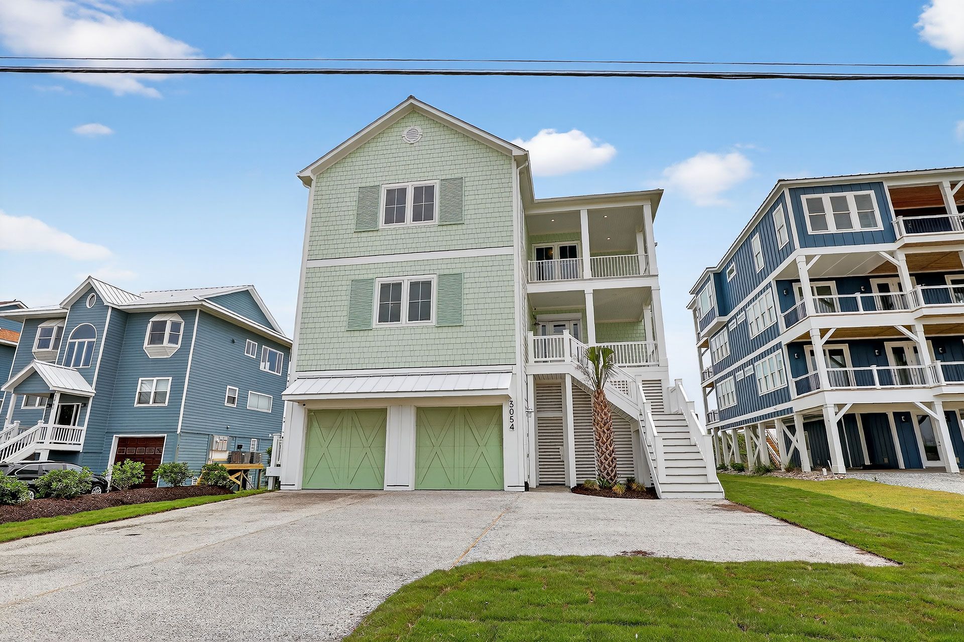 Three-story green beach house with two garage doors, balcony, and stairwell, flanked by blue houses, on a gravel driveway under a blue sky.
