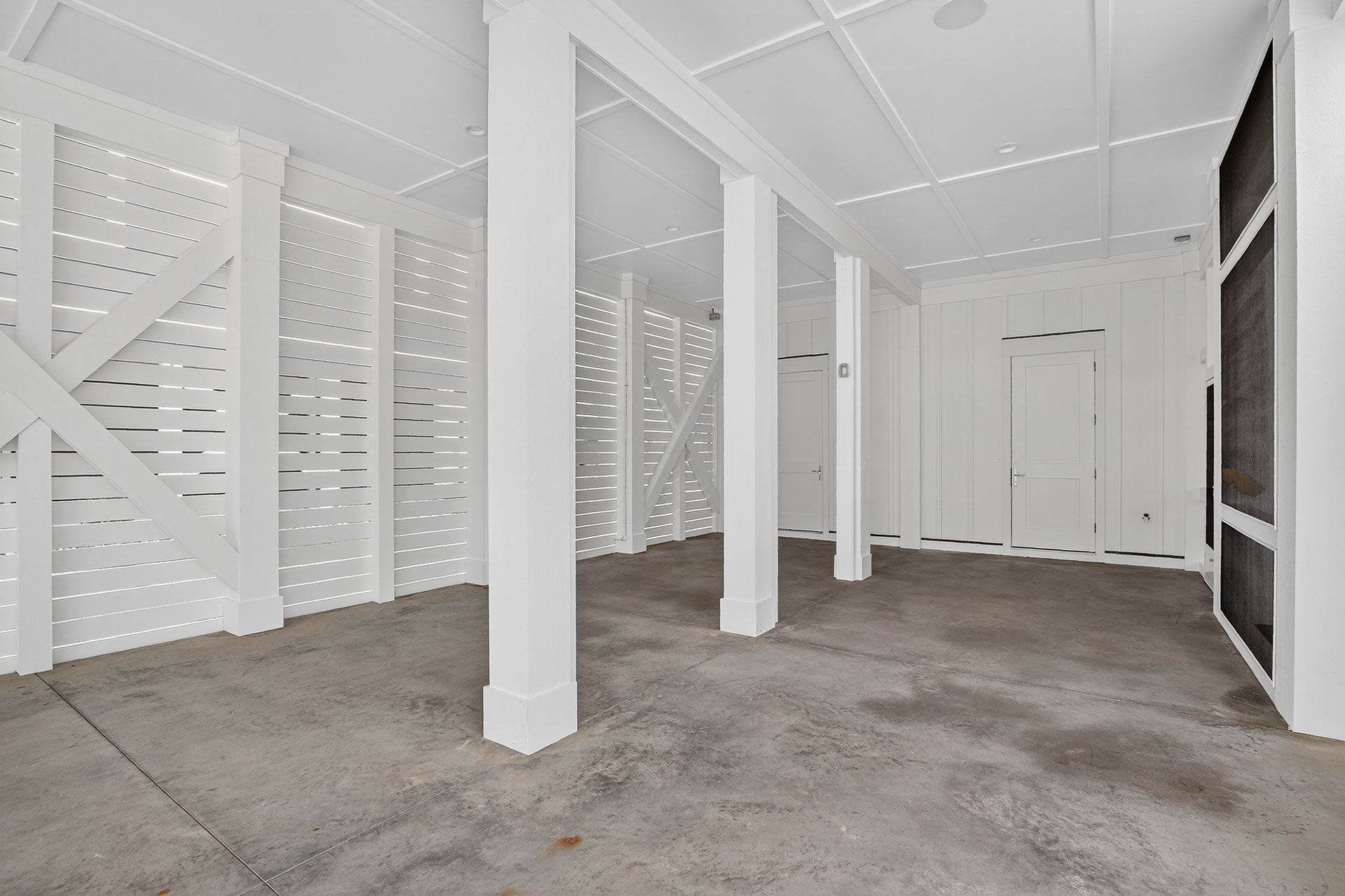 White-walled, empty room with concrete floor, supported by white columns. Wooden lattice walls, and a black screen.