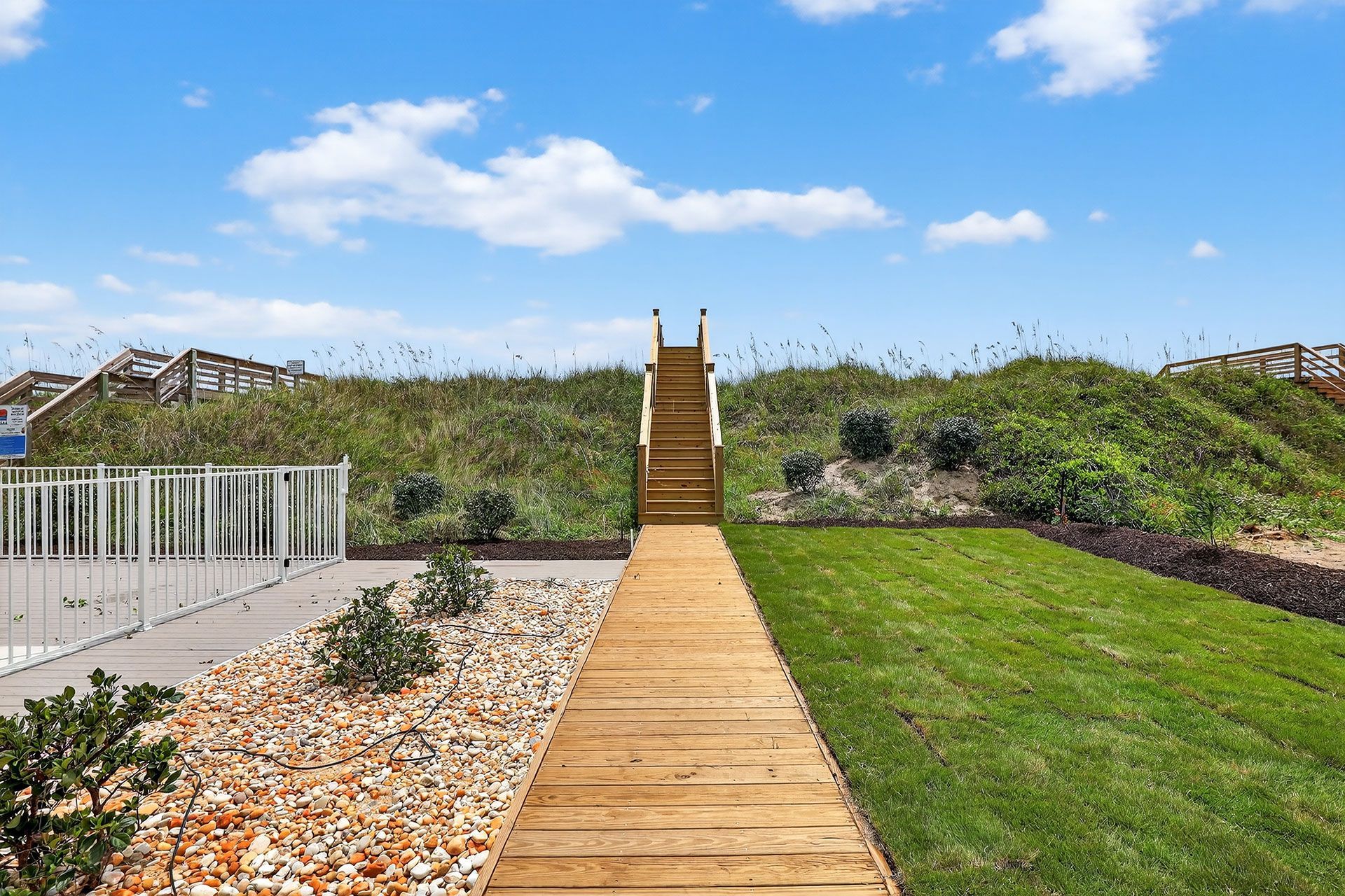 Wooden walkway and stairs leading to the beach over a grassy dune under a blue sky.