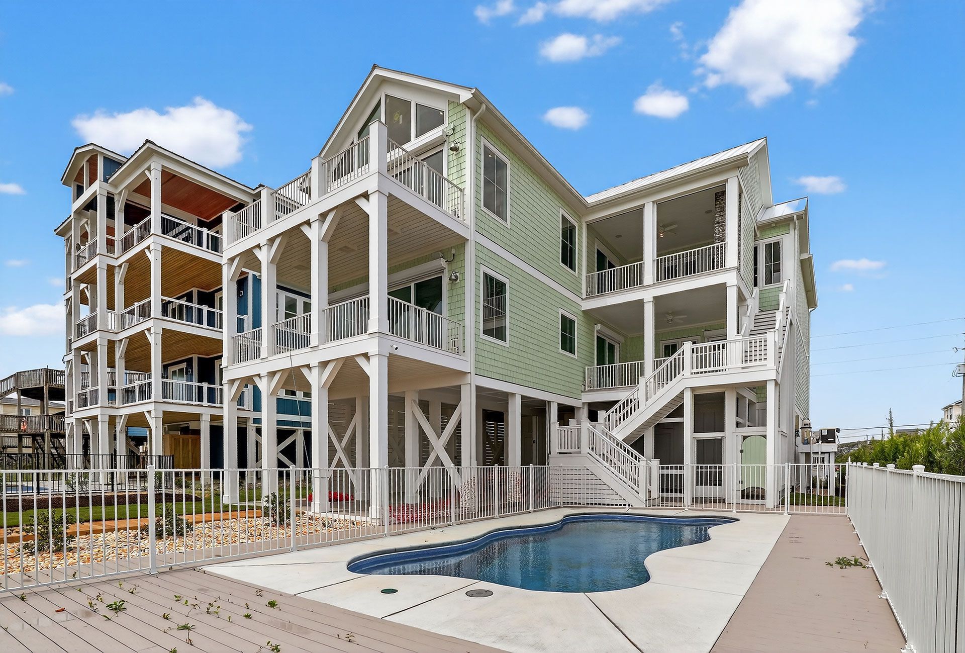 Beach house with multiple levels, balconies, pool, white and green exterior, blue sky.