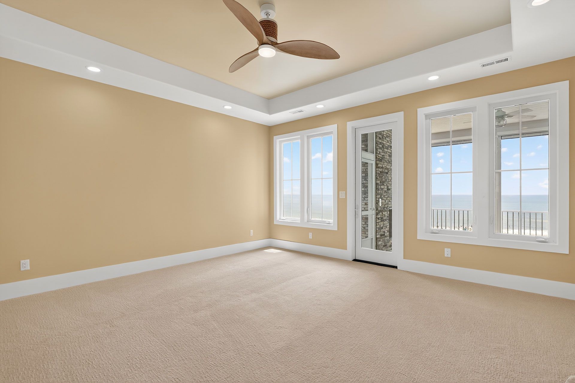 Empty bedroom with tan walls, ceiling fan, windows, and beige carpet.