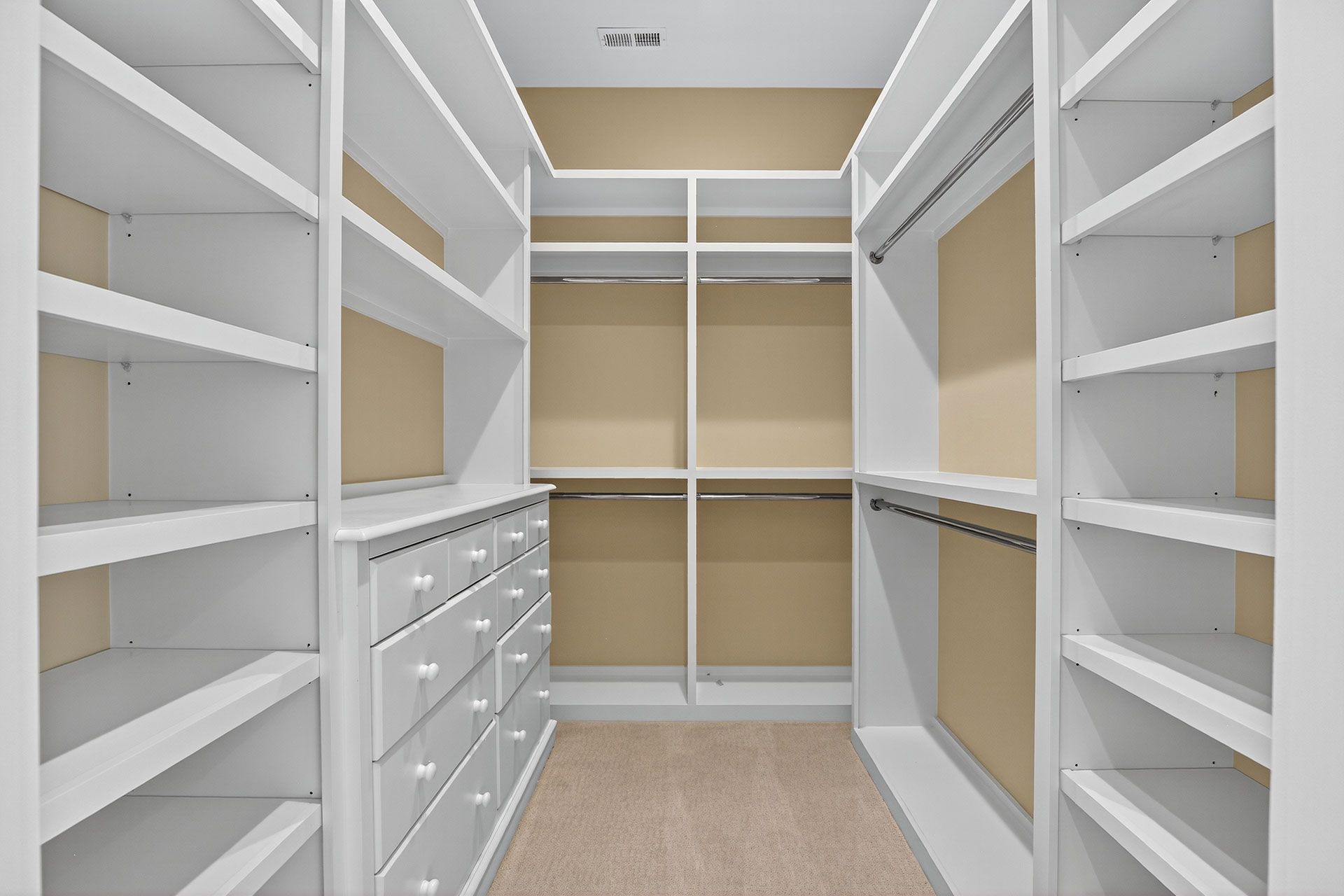 Empty, white walk-in closet with shelves, drawers, and hanging rods. Beige carpet and walls.