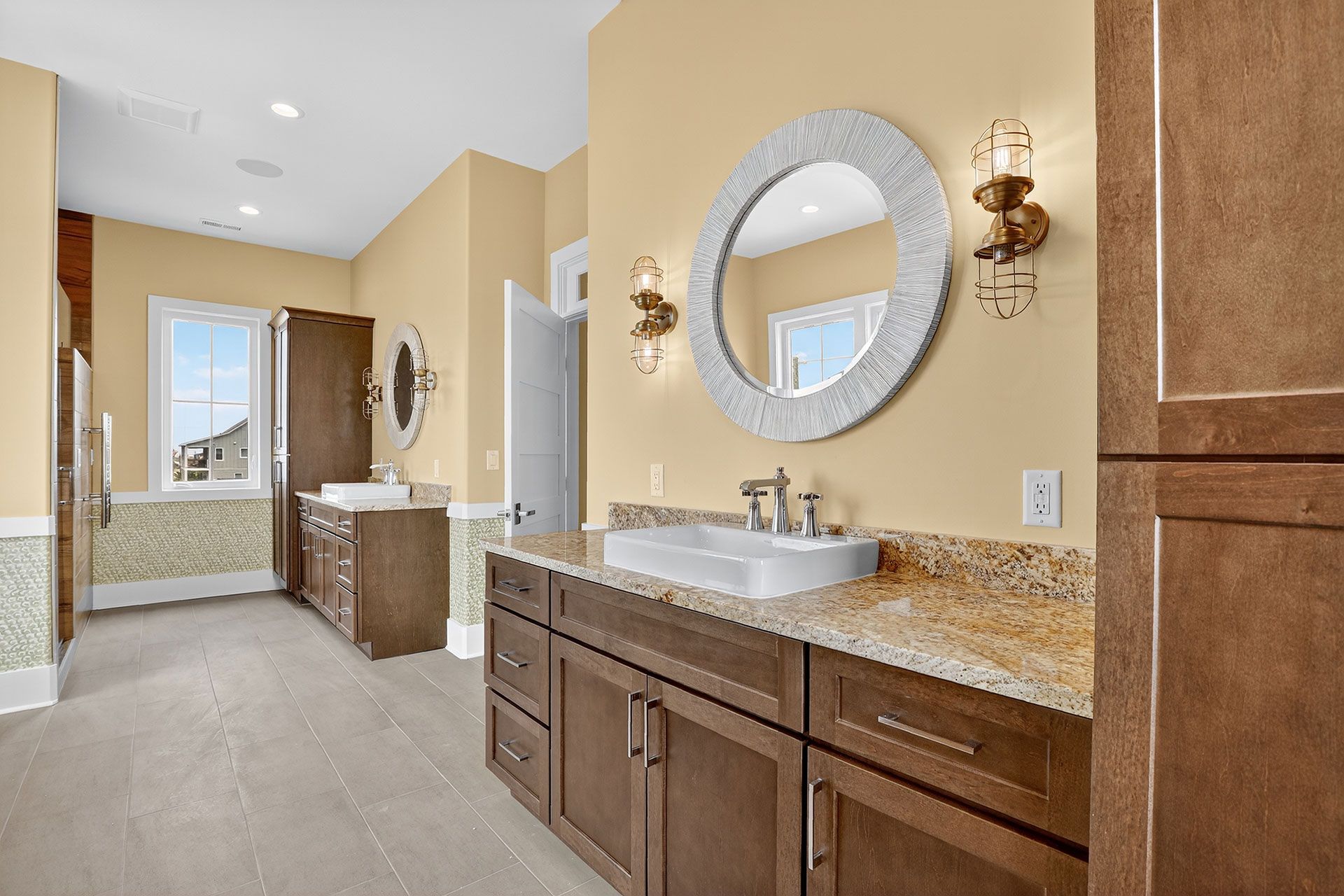 Bathroom with brown cabinets, tan countertops, large mirrors, sconces, and beige walls.