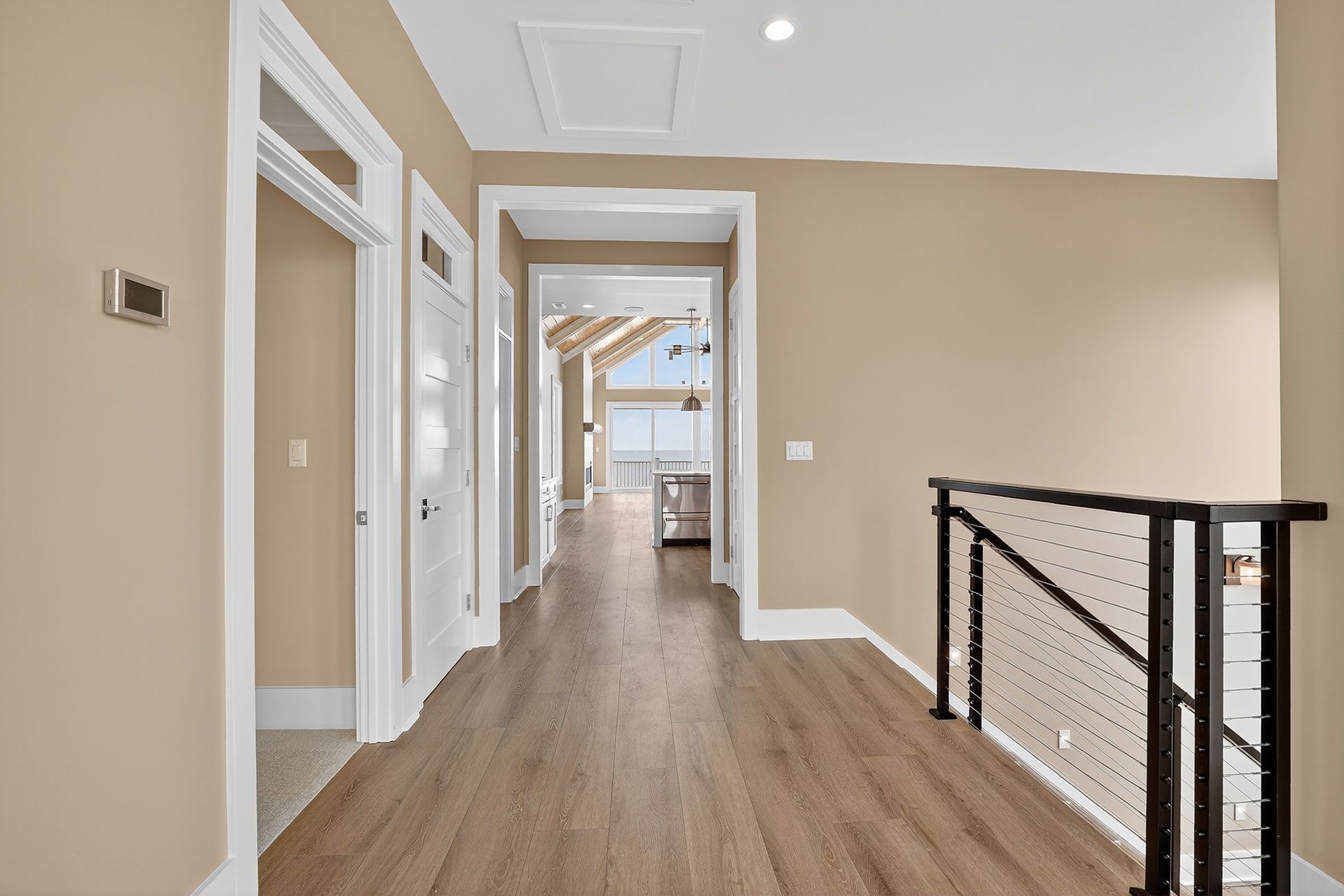 Hallway with light brown walls and flooring, white trim, and a black and silver railing.