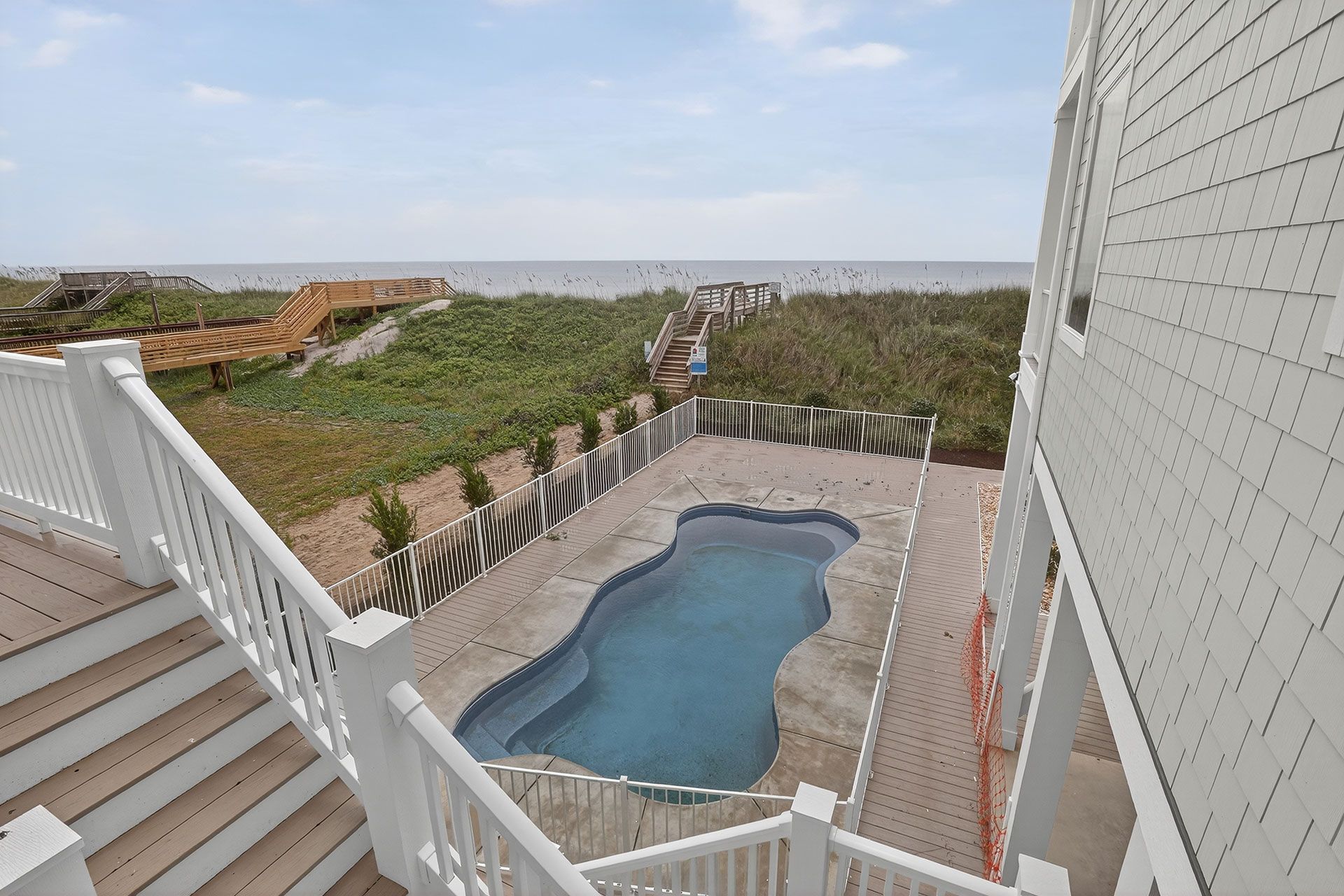 Beach house with a pool overlooking the ocean; blue sky, white railings, brown deck, and green vegetation.