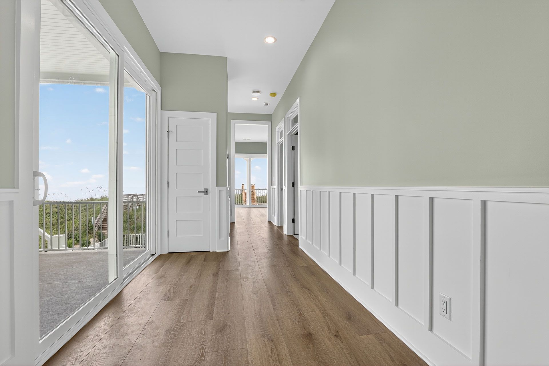 Hallway with light wood floors, white wainscoting, sage green walls, and sliding glass doors to a balcony.