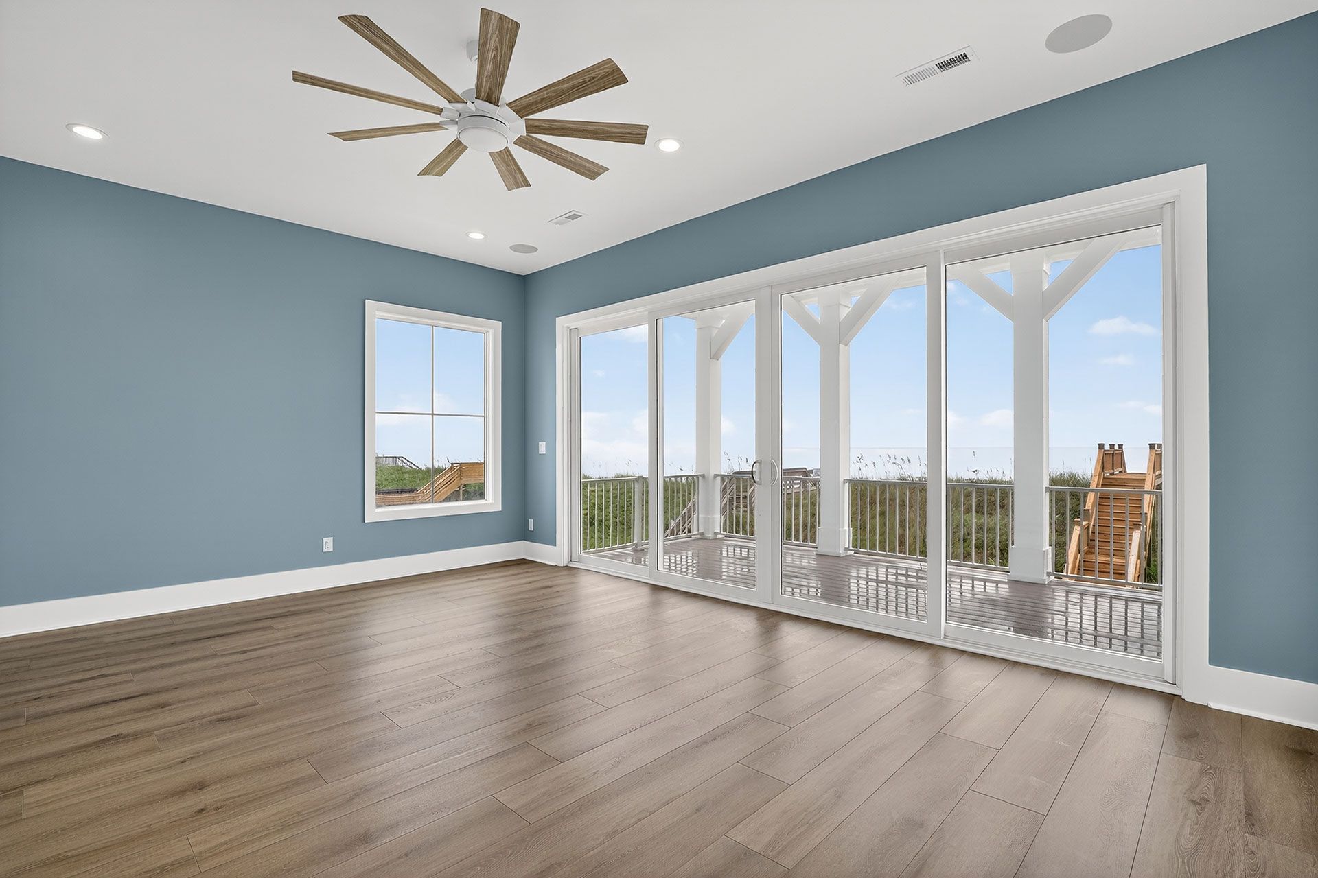 Empty room with blue walls, wood floor, large windows, and ceiling fan.