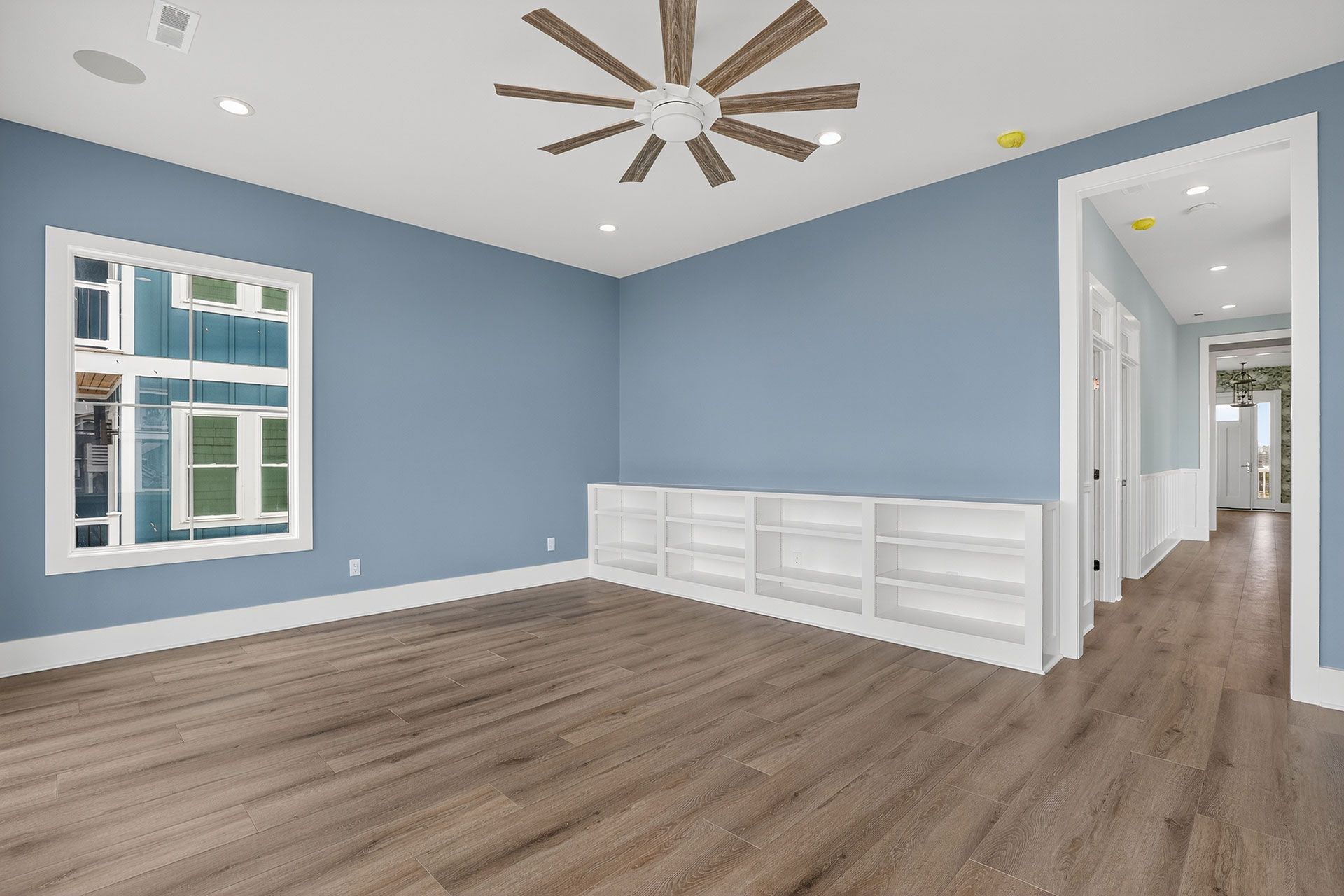 Empty room with blue walls, built-in white cabinets, wood floor, window, and ceiling fan.